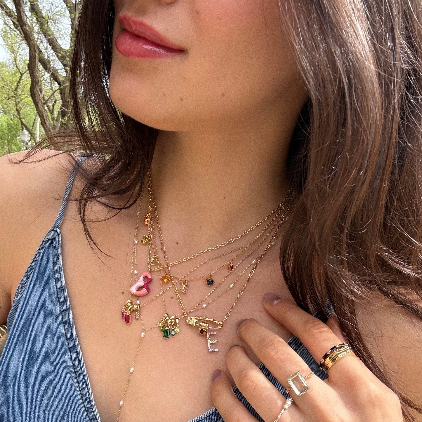 A woman poses outdoors in sunlight wearing a denim top, layered gold necklaces, colorful rings, and the Solid Gold Pastel Gemstone Letter Charm, touching her chest with manicured nails against a backdrop of trees.