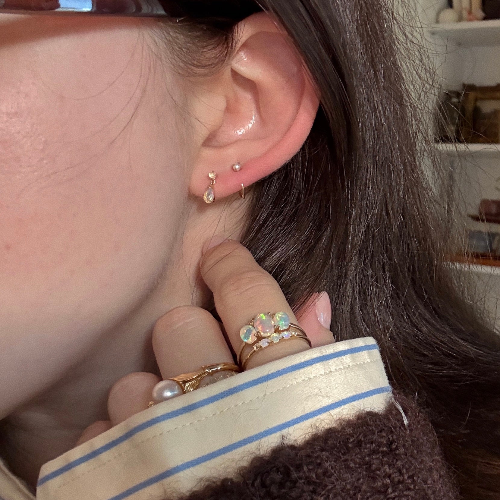 A close-up of a person’s ear shows the Solid Gold 2025 October Capsule Delicate Opal Drop Earrings alongside two small studs. Their hand, adorned with gold rings accented by color and pearls, rests near brown hair and a striped sleeve.
