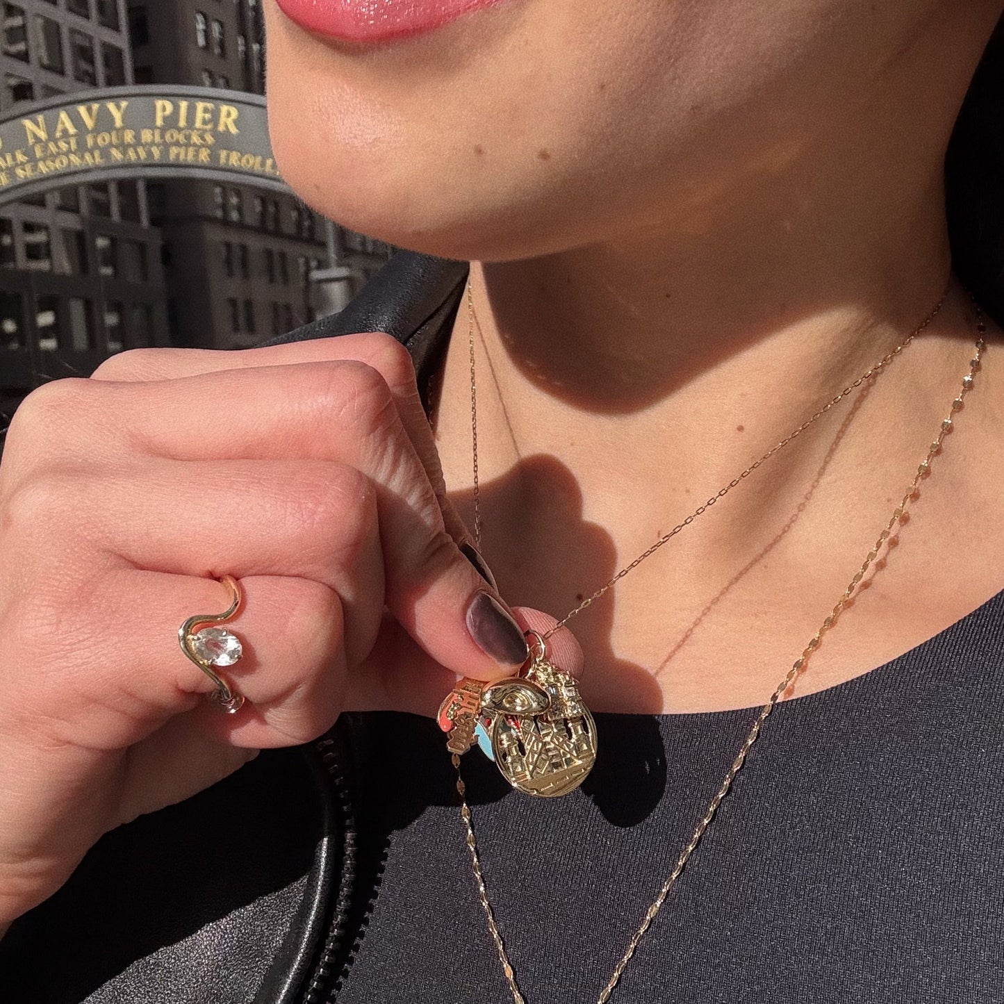 A person in a dark top and leather jacket holds a Solid Gold Chicago Charm necklace; a matching ring with a clear stone is on their finger. In the background, a “Navy Pier” sign and city buildings are visible.