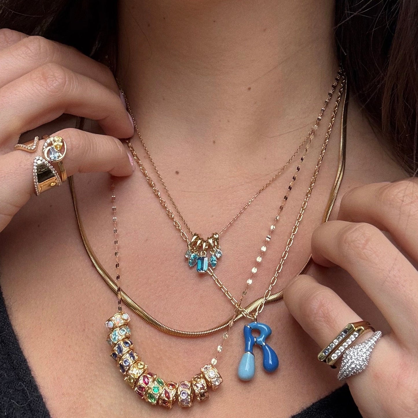 A person wearing layered gold necklaces with colorful charms and pendants, including a gold-plated charm and jeweled beads, plus rings adorned with gems, diamonds, and the Shades of You Birthstone Spacer Charm on both hands.
