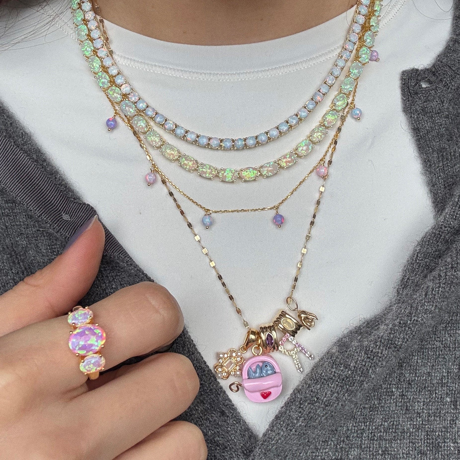 A person wears layered pastel gemstone necklaces, one with gold charms including a pink Tamagotchi and MOM letters. They also wear the Pink Opal Gum Drop Ring with large iridescent stones and a gray jacket over a white shirt.