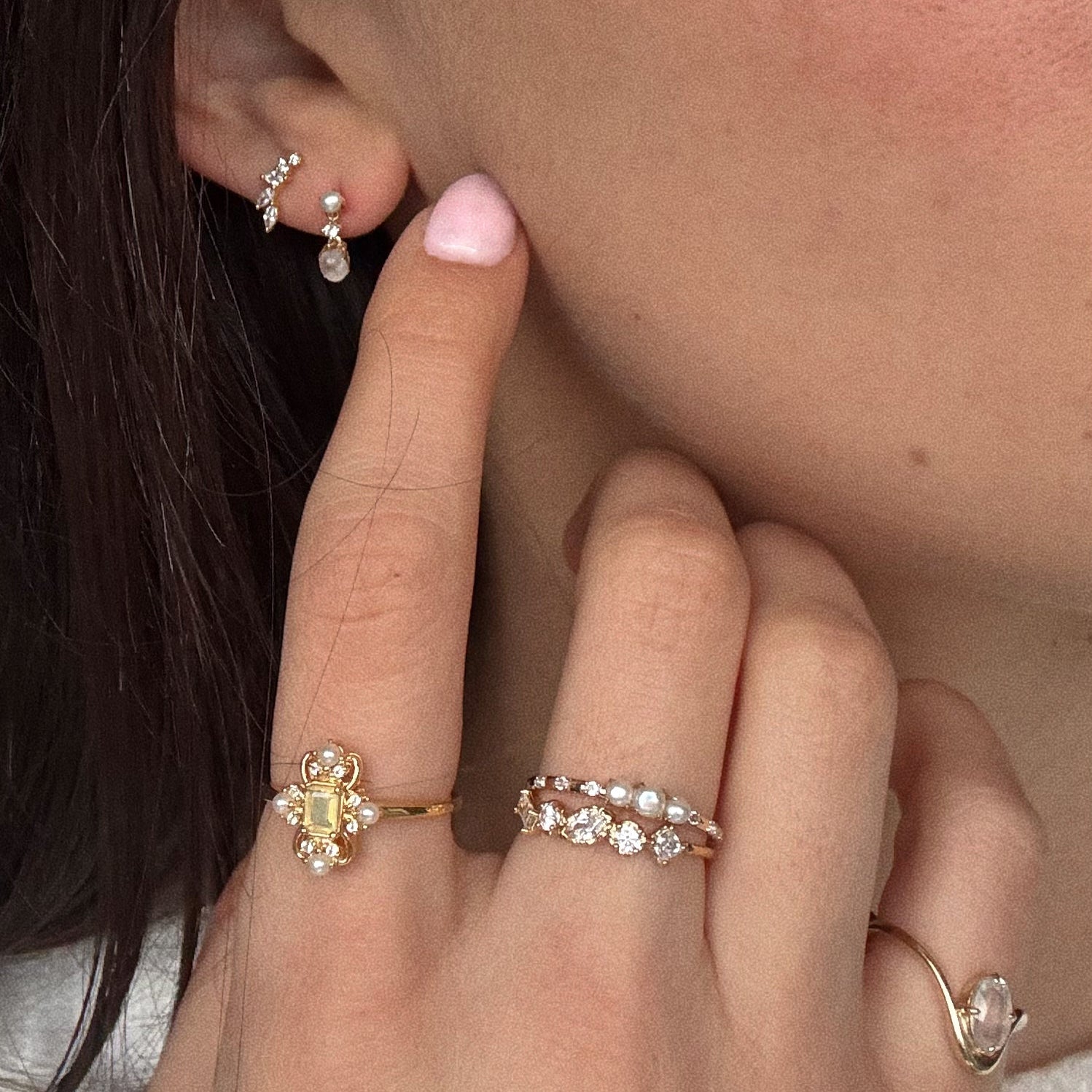 A close-up shows a person’s ear and hand adorned with gold rings featuring pearls and gemstones, paired with Solid Gold Soft Glow Pearl and Moonstone Drop Studs. Their nails are painted light pink.