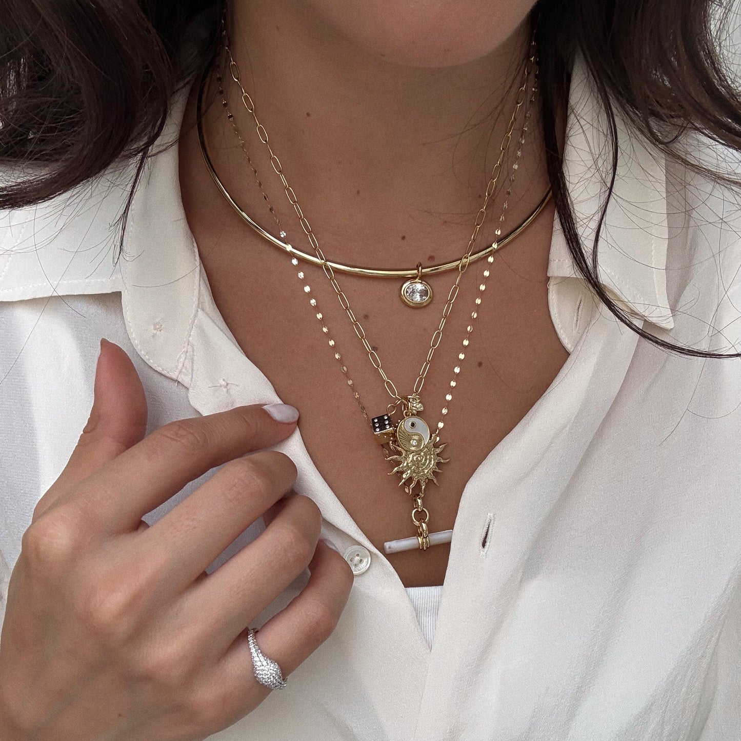 A woman in a white blouse showcases Charms by Mod + Jo, featuring layered gold-plated pendants like a sun, pearl, and crystal—believed by some to bring good luck—plus a silver ring on her finger.