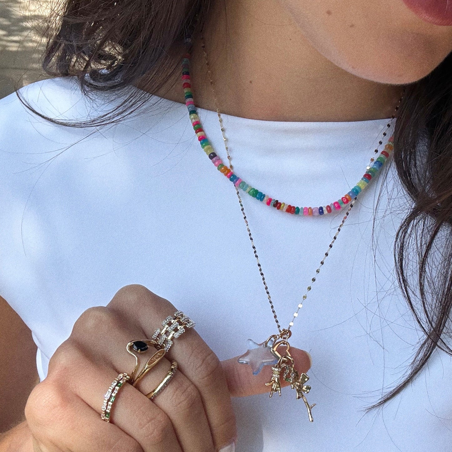 A person in a white top and colorful beaded necklace wears the Luiny x Local Eclectic Charms gold chain. Their hand, decorated with several gold rings, gently touches the necklaces.