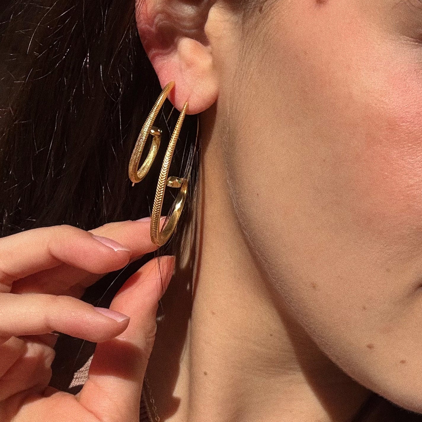Close-up of a person’s ear wearing two Mini Snake Hoop Earrings, plated in 18k gold. Their hand gently touches one hoop as sunlight highlights the earrings and skin, drawing attention to the jewelry and ear.