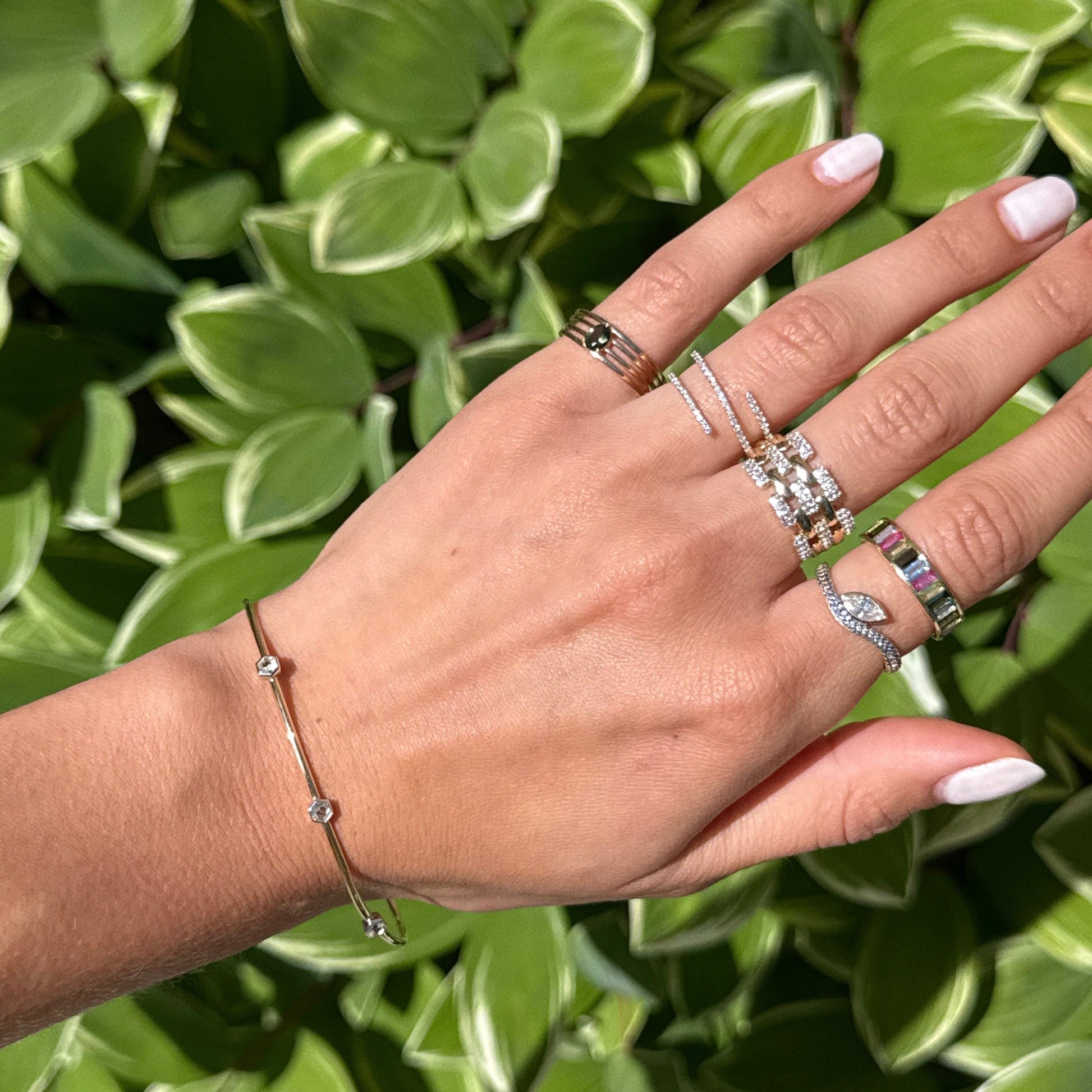 A hand with manicured nails showcases several gold and silver rings with diamonds and white topaz, along with a Solid Gold Geometric Glimmer Mixed Metal Bracelet, set against a backdrop of green leaves.