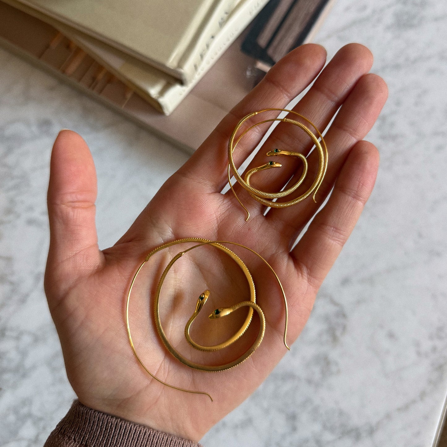 A hand holds two Mini Snake Hoop Earrings, 18k gold plated and coiled in spirals with green zircon eyes. Books and a marble surface can be seen in the background.