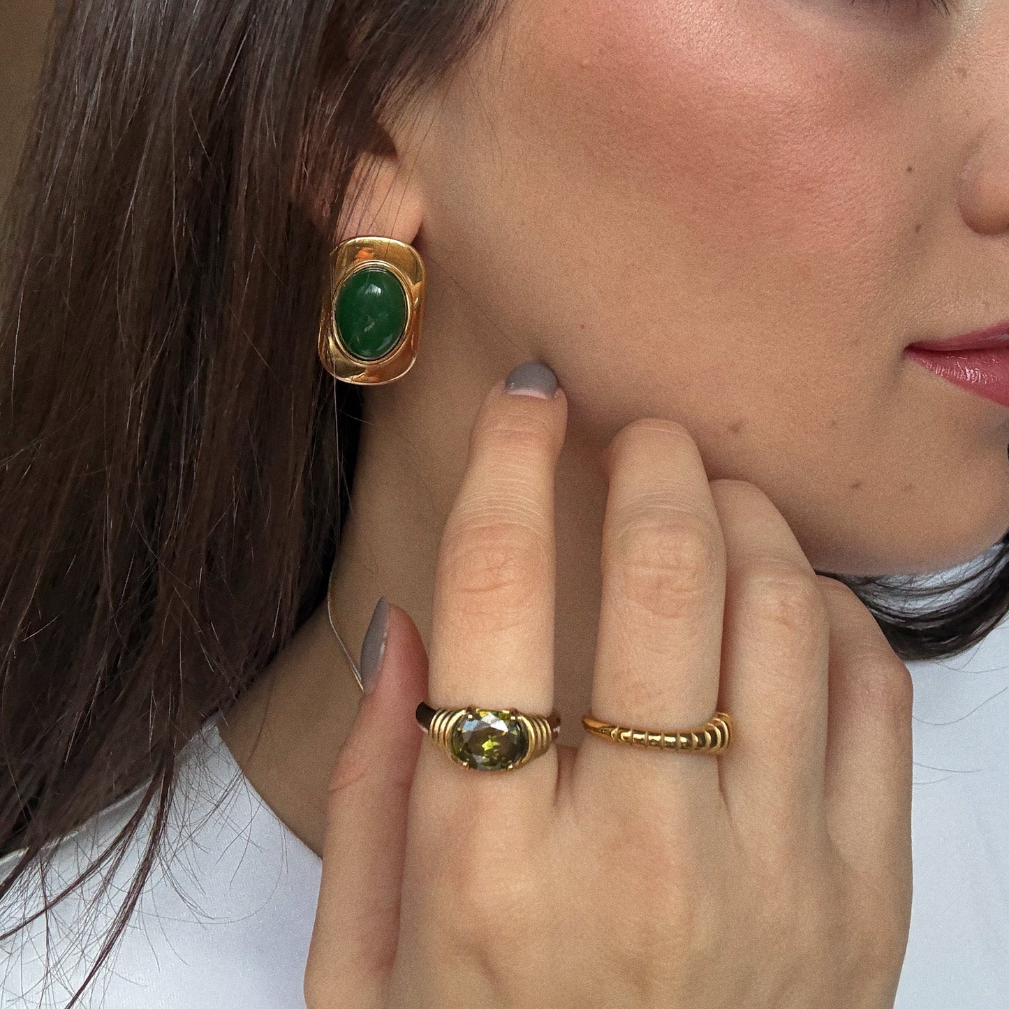 A close-up of a woman with straight brown hair touching her cheek, wearing gold earrings with green stones, the Stone Statement Ring featuring a green gemstone, and a simple gold band.