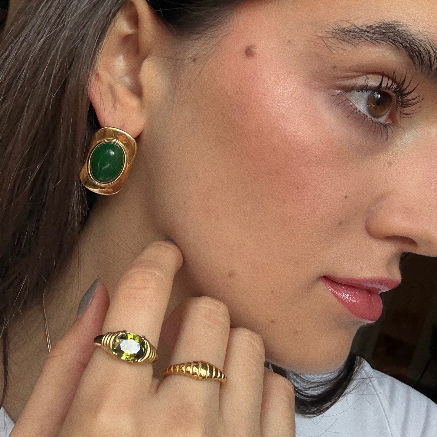 Close-up of a woman with smooth skin and brown hair, wearing natural makeup, a green stone earring, the 18k gold plated Crew Ring by Petit Moments with a green gemstone, and a sleek gold band.