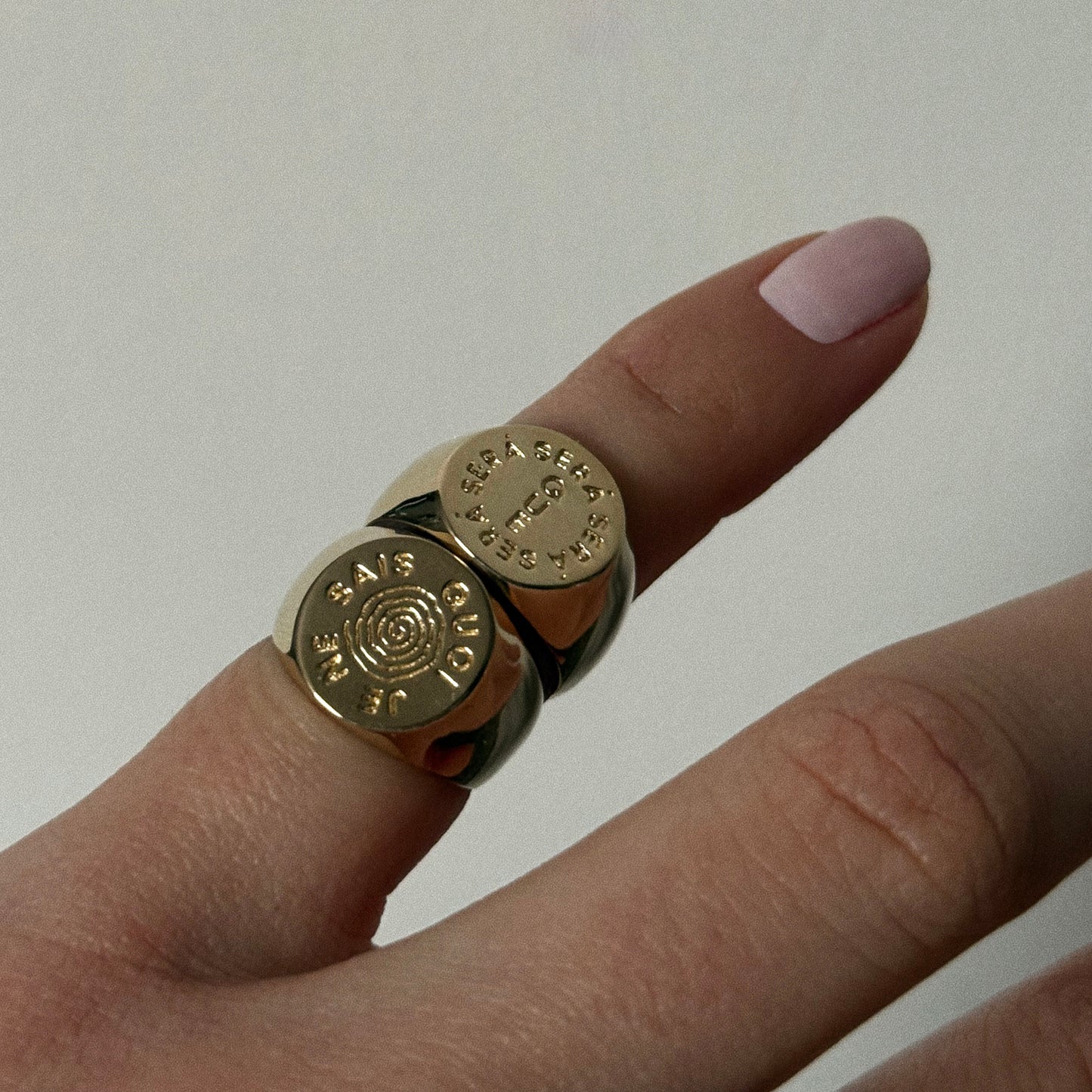 A hand with light pink nail polish wears the I Like It Here Club x Local Eclectic Je Ne Sais Quoi Pinky Ring, a chunky gold piece engraved with circular patterns and THE SUN SAYS YES. The background is plain and neutral.