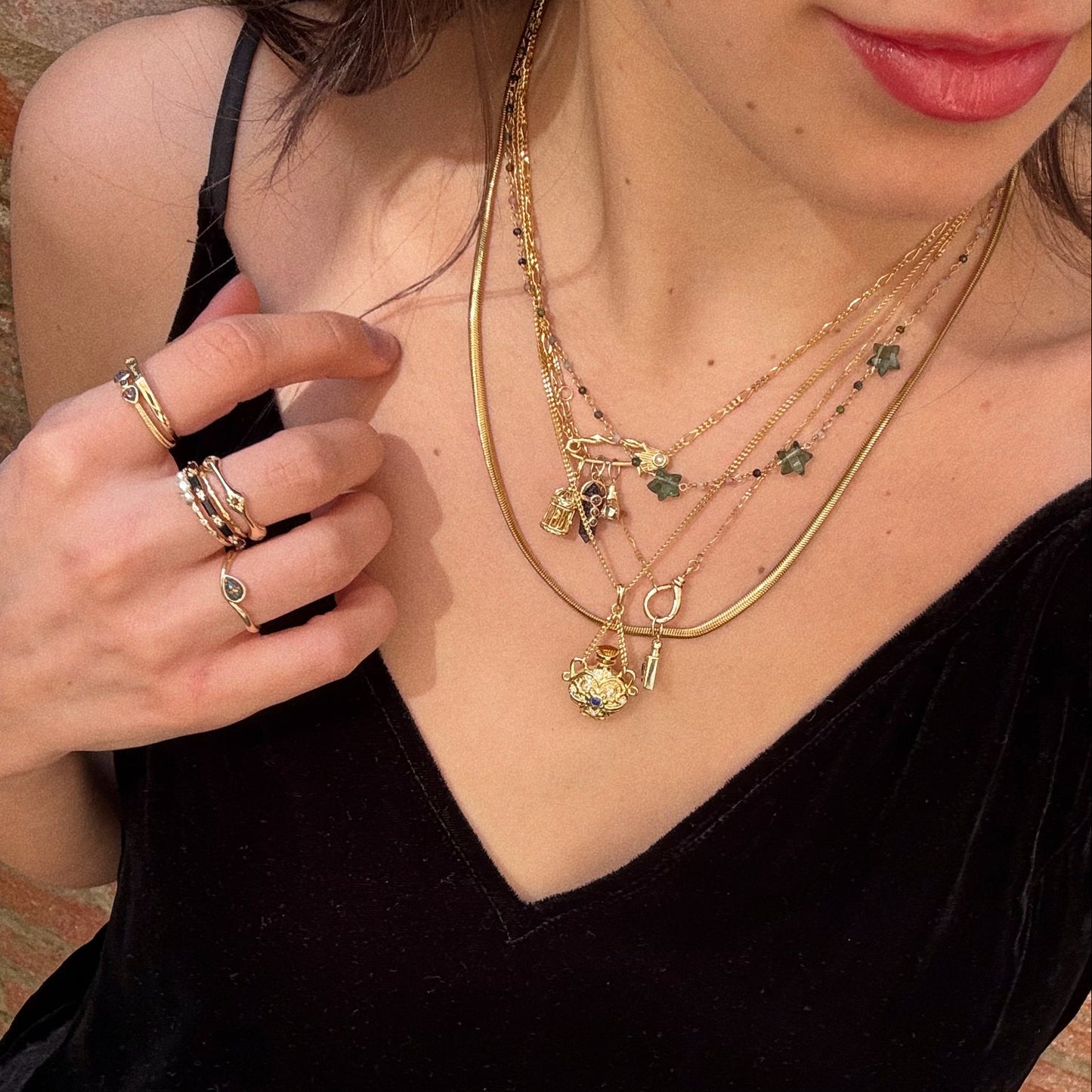 A woman in a black V-neck top wears multiple layered necklaces, including the Classic Snake Chain Necklace adorned with charms and pendants. She also wears gold rings, her lips slightly parted as her hand touches her shoulder.