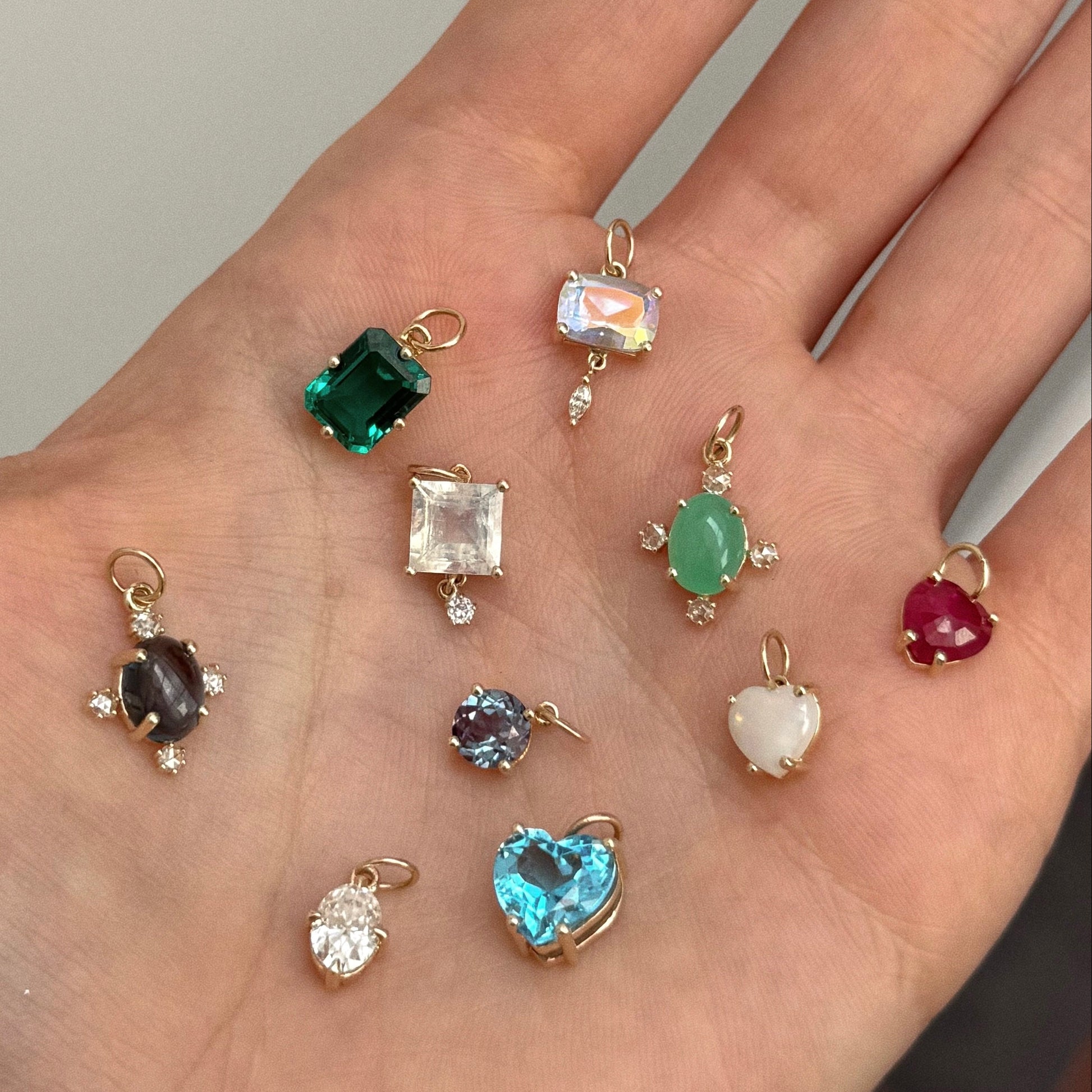 A hand holds ten One of a Kind 14k Chrysoprase and Diamond Charms—solid yellow gold pendants set with cabochon chrysoprase, rose cut diamond, and various gemstones in heart, oval, and square shapes with colorful hues.