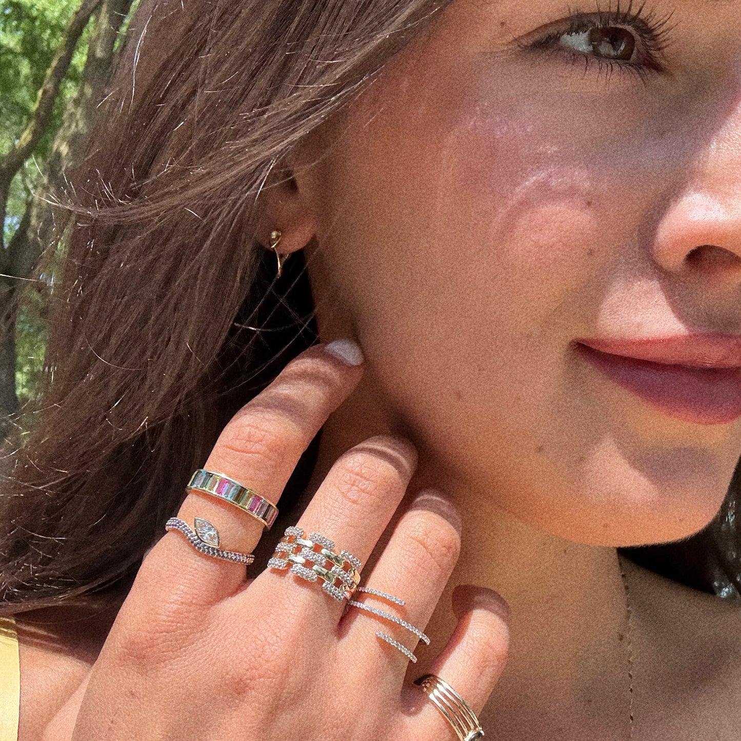 A close-up of a woman’s face and hand wearing the Solid Gold Nesting Gleam Sapphire and Diamond Ring, sunlight accentuates her jewelry and hair, with soft green foliage blurred in the background.