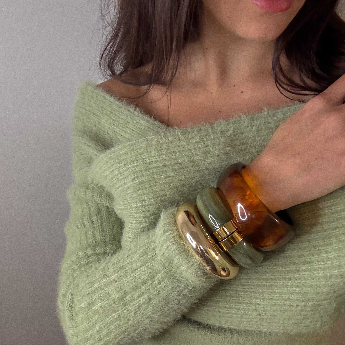 A woman, her hair loose and lower face shown, wears a pale green off-shoulder sweater. On her wrist are Bangle Bracelets: shiny gold plated, translucent amber, and sage green marbled resin.