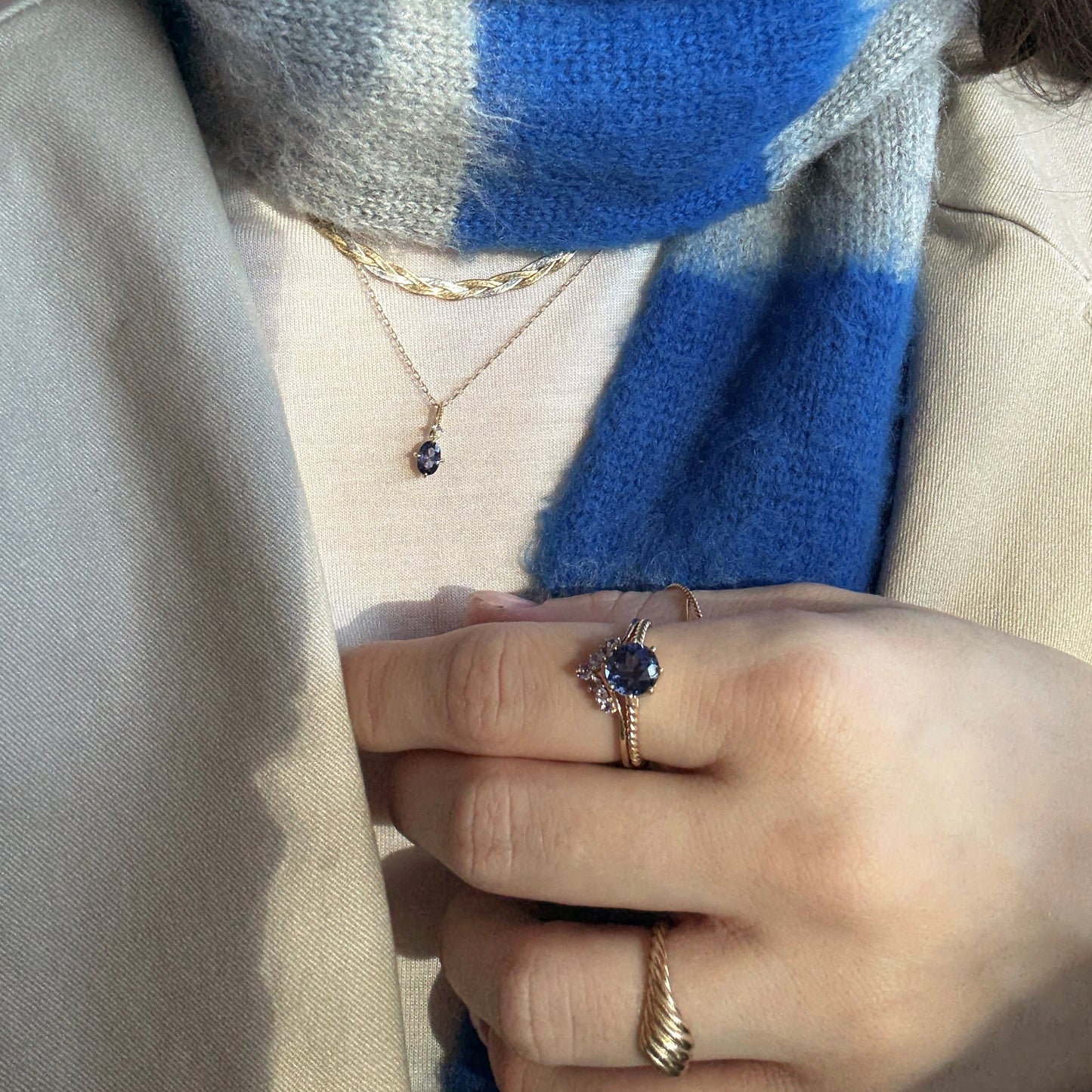 A person wears a blue and gray scarf, beige coat, and the Solid Gold 2025 December Capsule Classic Tanzanite Pendant Necklace. Their hand displays gold rings, one with a blue gemstone and another with a twisted design.