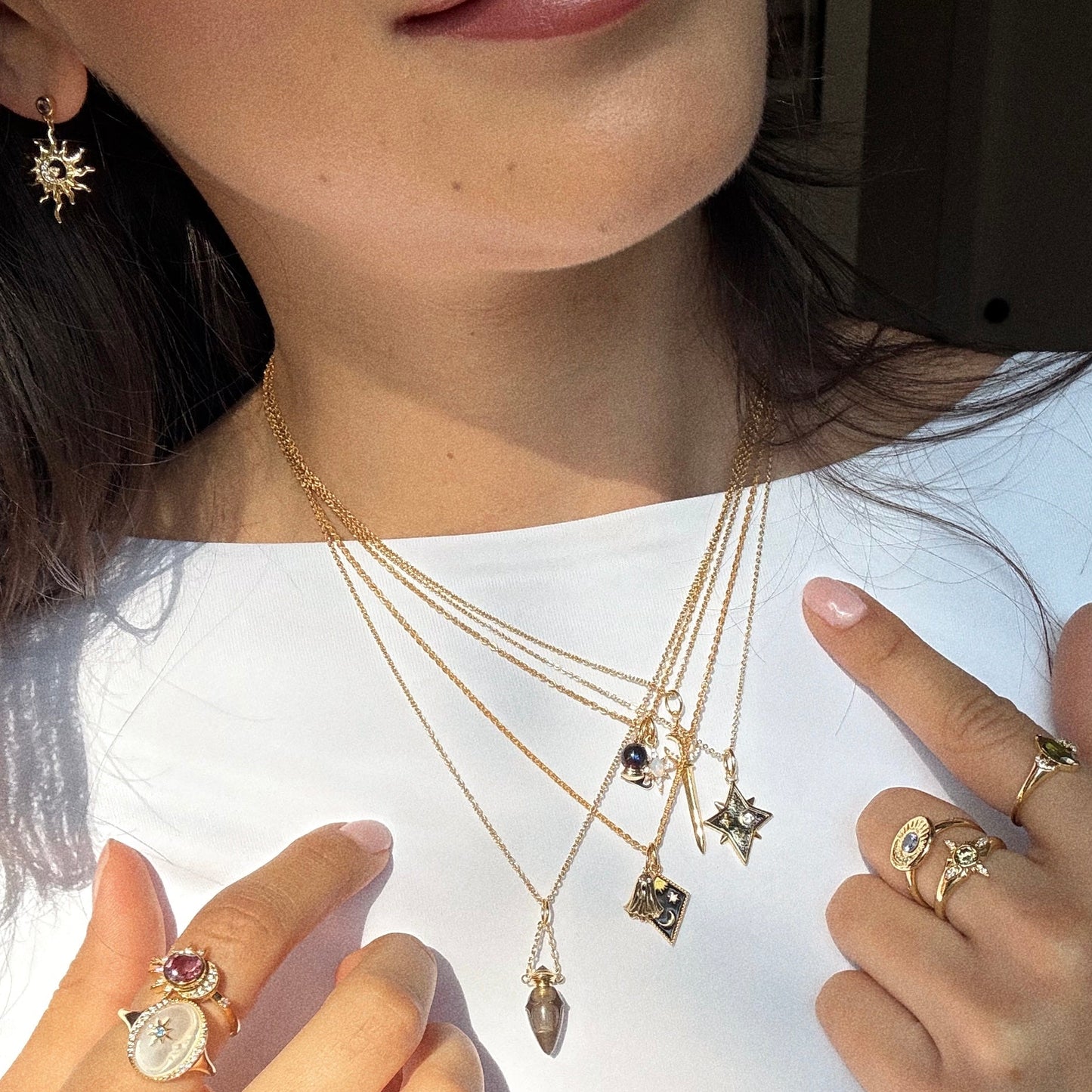 A woman in a white top models mystical jewelry: layered gold necklaces, gold rings with colorful stones, a sun-shaped earring, and she gently holds two necklaces—one featuring the Solid Gold Superstition Charm.