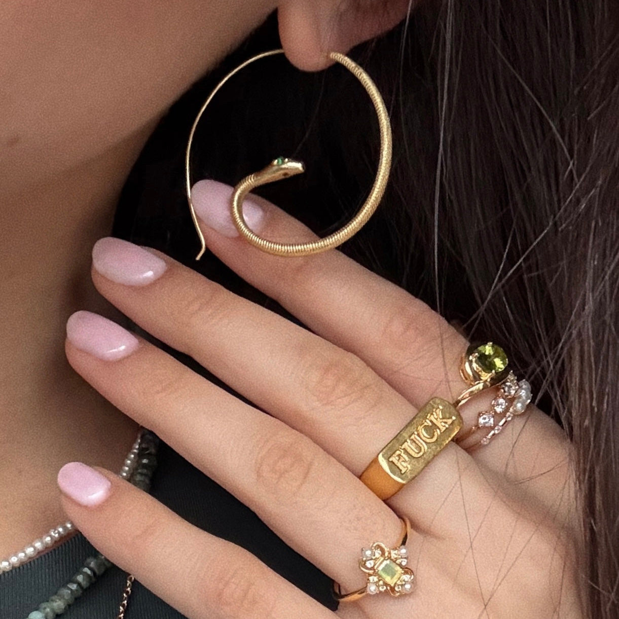 A person with light pink manicured nails wears striking Mini Snake Hoop Earrings and several rings, including a bold gold FUCK ring, a yellow gemstone ring, and delicate rings with pearls or green zircon stones.