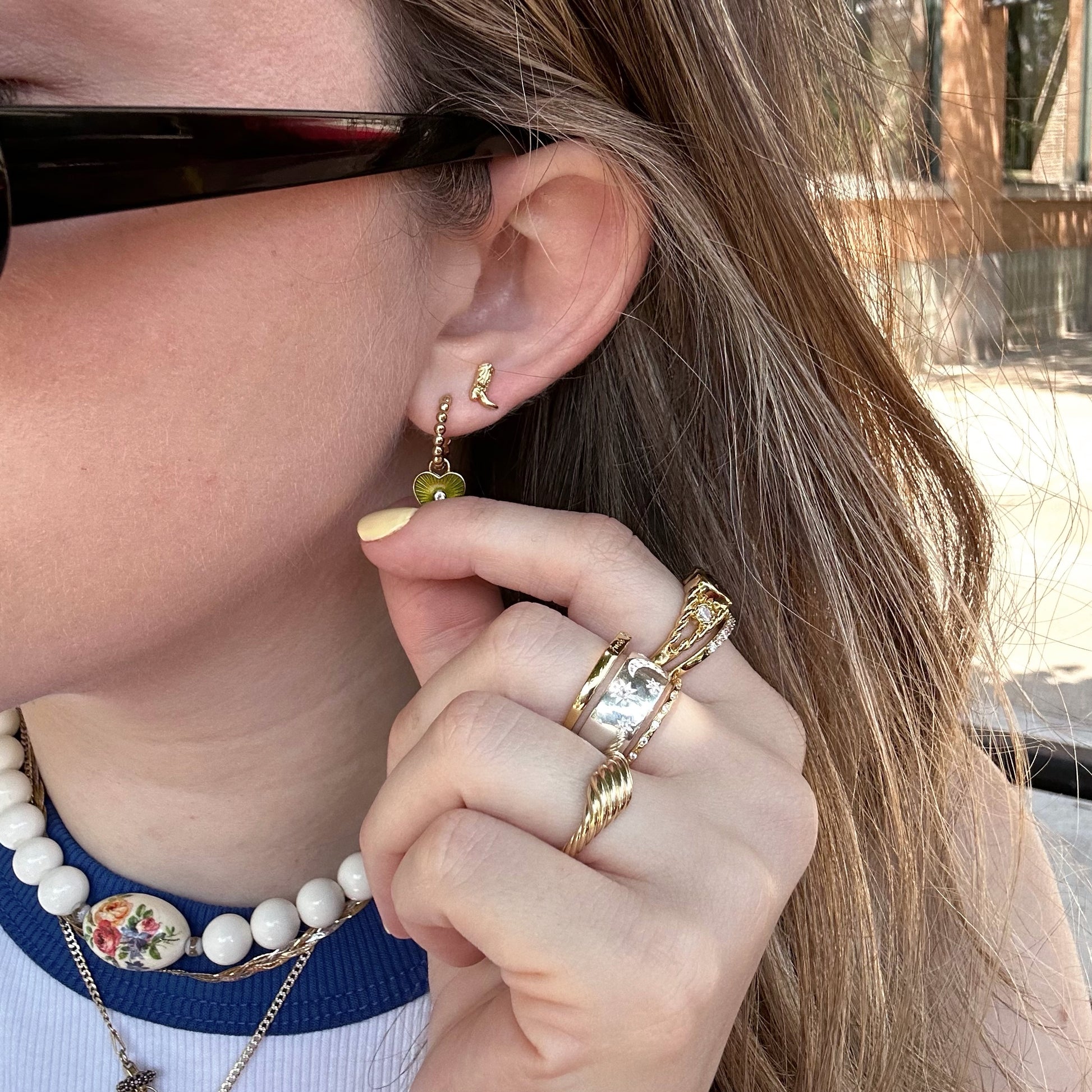 A person wearing black sunglasses, gold rings, a pearl necklace with a floral pendant, and 14k gold plated Cowboy Boot Studs holds their hand near their ear.