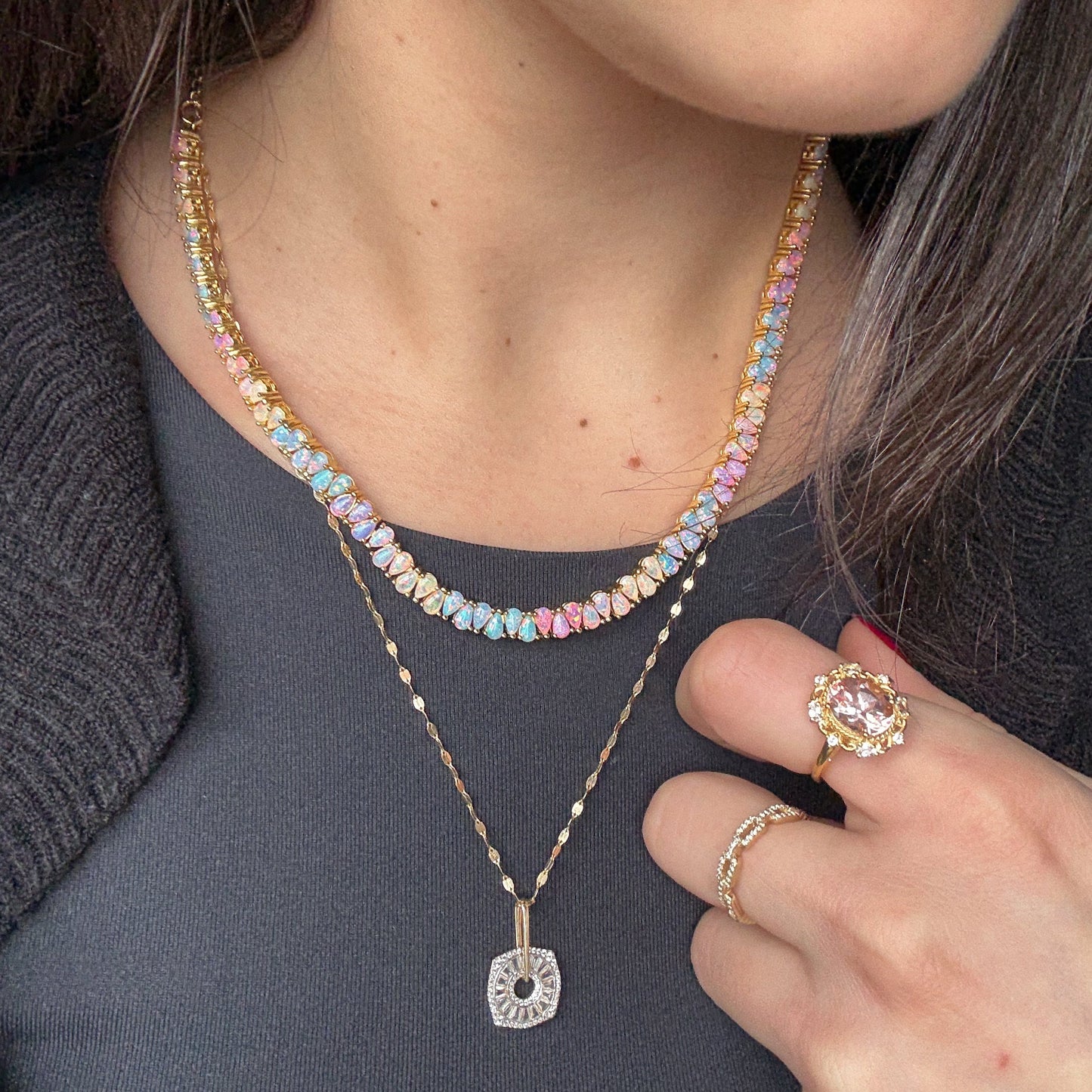 A woman in a dark top layers gold necklaces, including the Opal Dancing Dreams Tennis Necklace in 18k yellow gold, and showcases elegant gold rings—such as a large gemstone ring—on her hand near her chest.