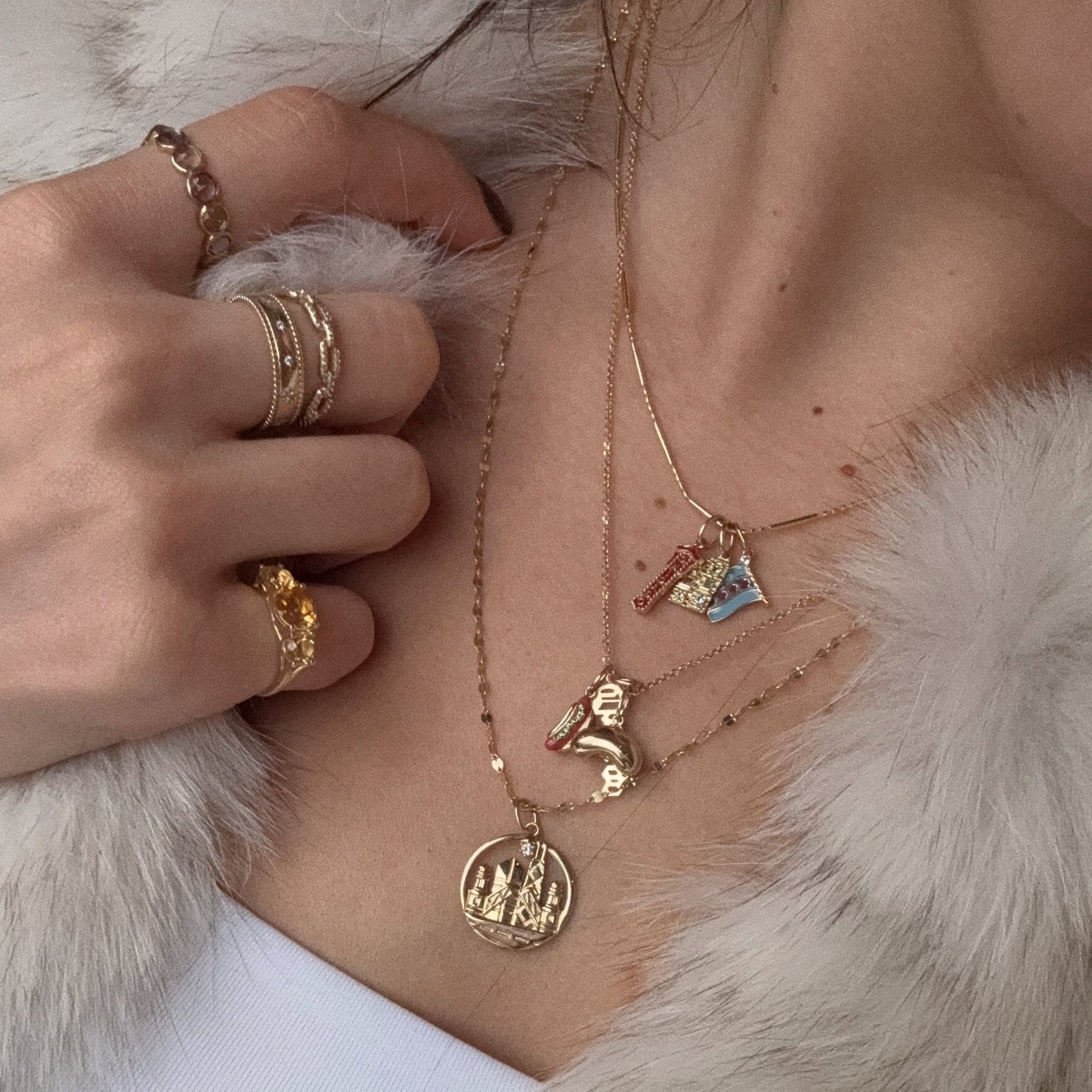 A close-up of a person wearing layered 9k solid yellow gold necklaces with pendants—a castle, letter "R," dinosaur, and mountain—plus a Solid Gold Chicago Charm and stacked gold rings, all paired with a light fur-textured jacket.