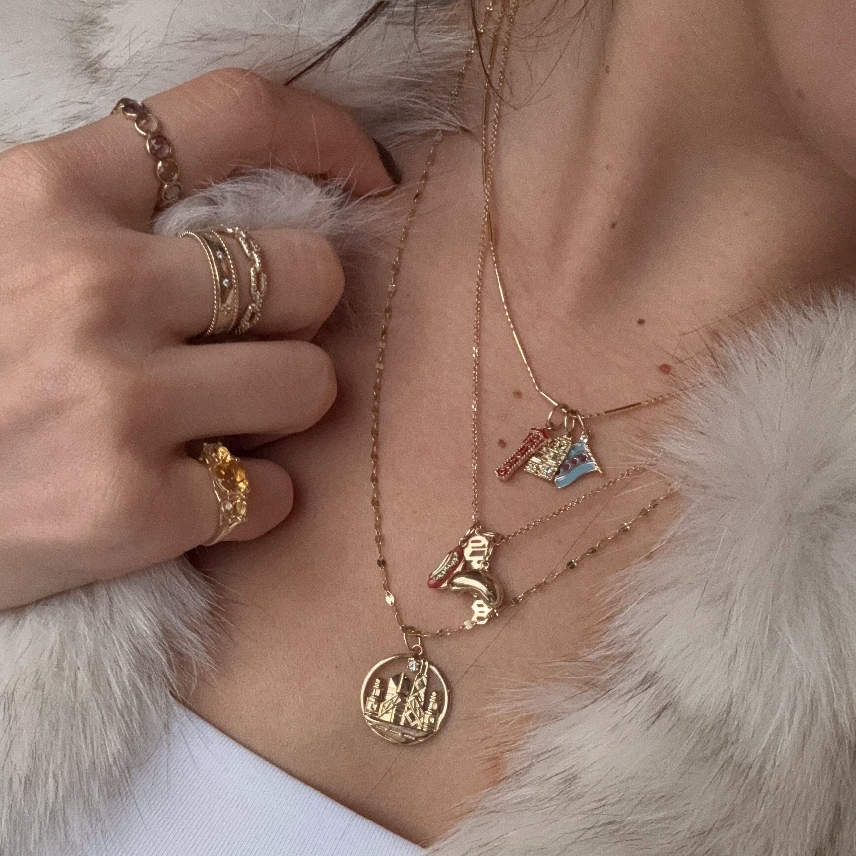 A close-up of a person wearing layered 9k solid yellow gold necklaces with pendants—a castle, letter "R," dinosaur, and mountain—plus a Solid Gold Chicago Charm and stacked gold rings, all paired with a light fur-textured jacket.