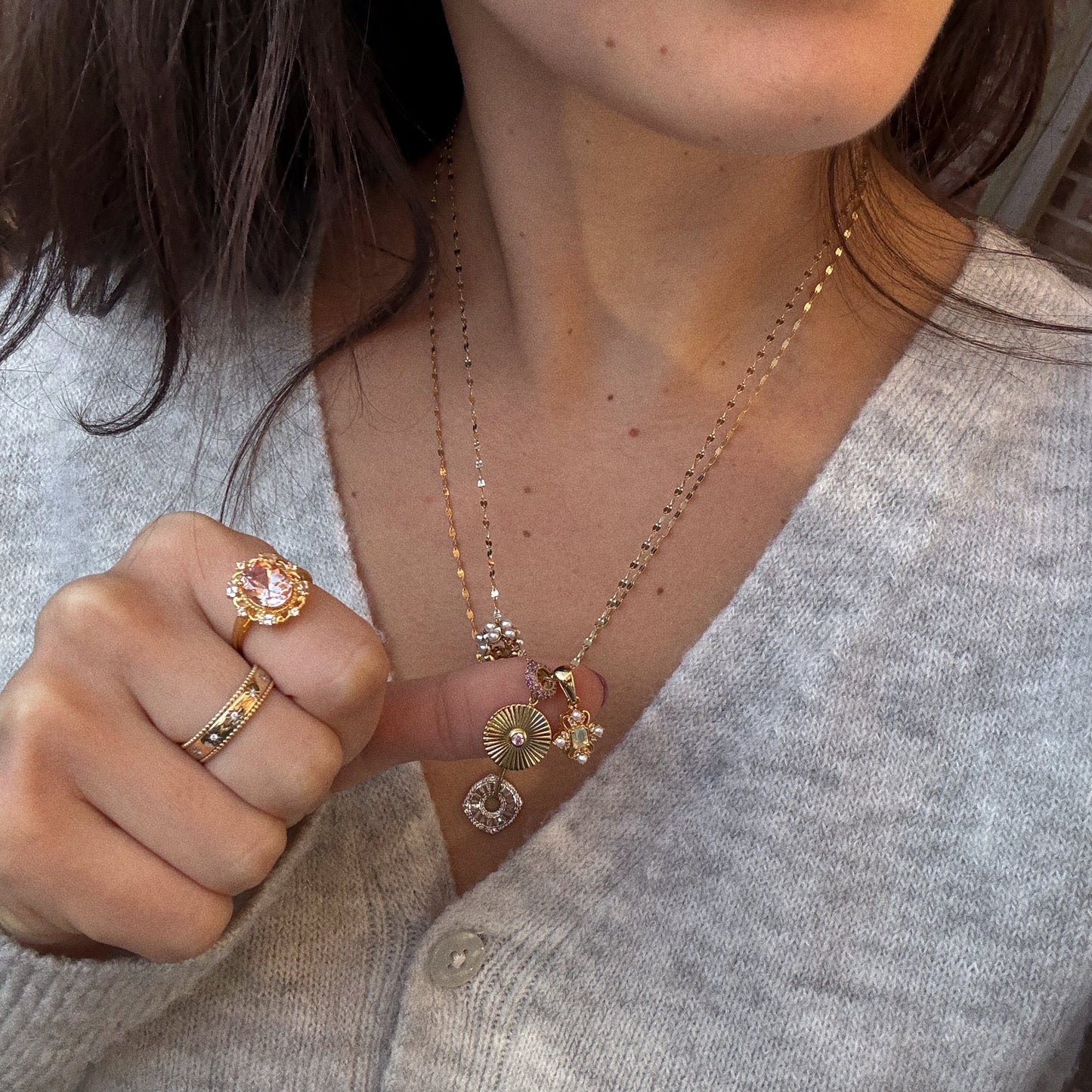 A woman in a light gray sweater displays gold rings, featuring the Royal Blush Cocktail Ring, and layered gold necklaces with decorative pendants. Her lower face and hand are visible as she holds her hand near her chest.