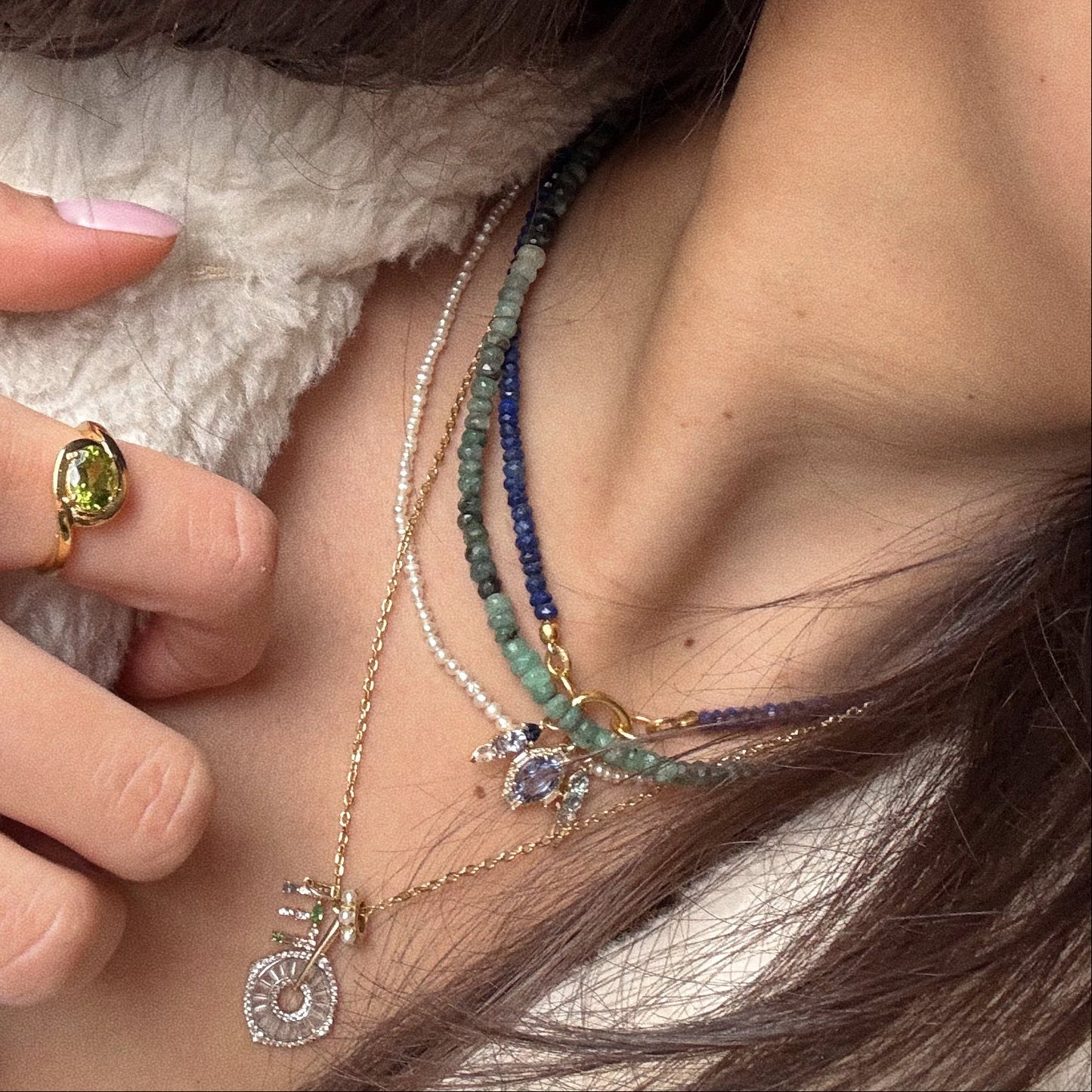 A close-up of someone wearing the Beaded Gemstone Charm Holder Necklace with colorful charms, including a sun-shaped charm holder, plus a gold ring with a green gemstone, while holding their necklace in a fuzzy white garment.