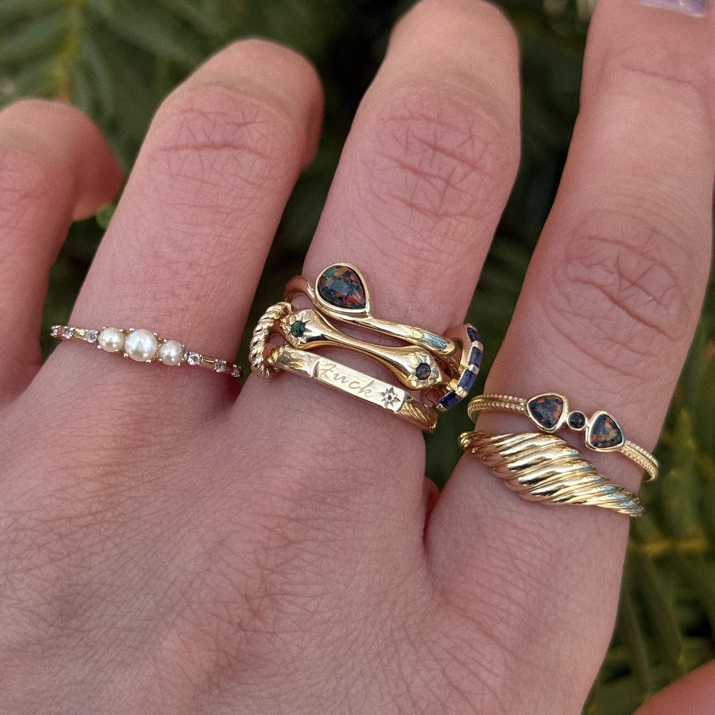 A hand adorned with the Solid Gold Tiny Fuck Ring and three other unique gold rings—featuring pearls, white topaz, engraved text, and twisted bands—is shown up close against lush green foliage.
