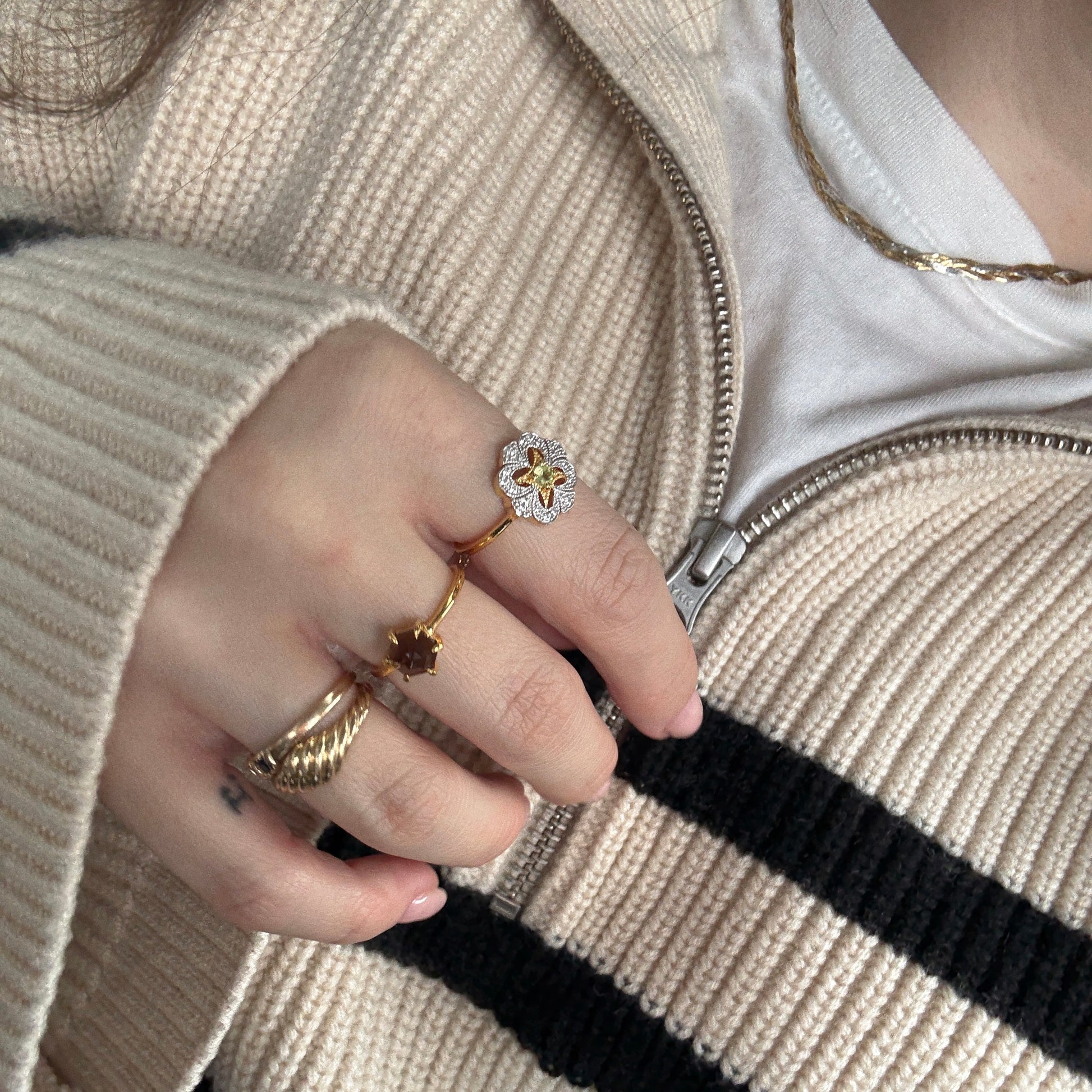 Someone wearing a cream and black striped zip-up sweater holds the collar, displaying vintage-style gold rings—including the Hexagon Smokey Quartz Ring—and a thin 14k gold vermeil necklace over a white shirt.