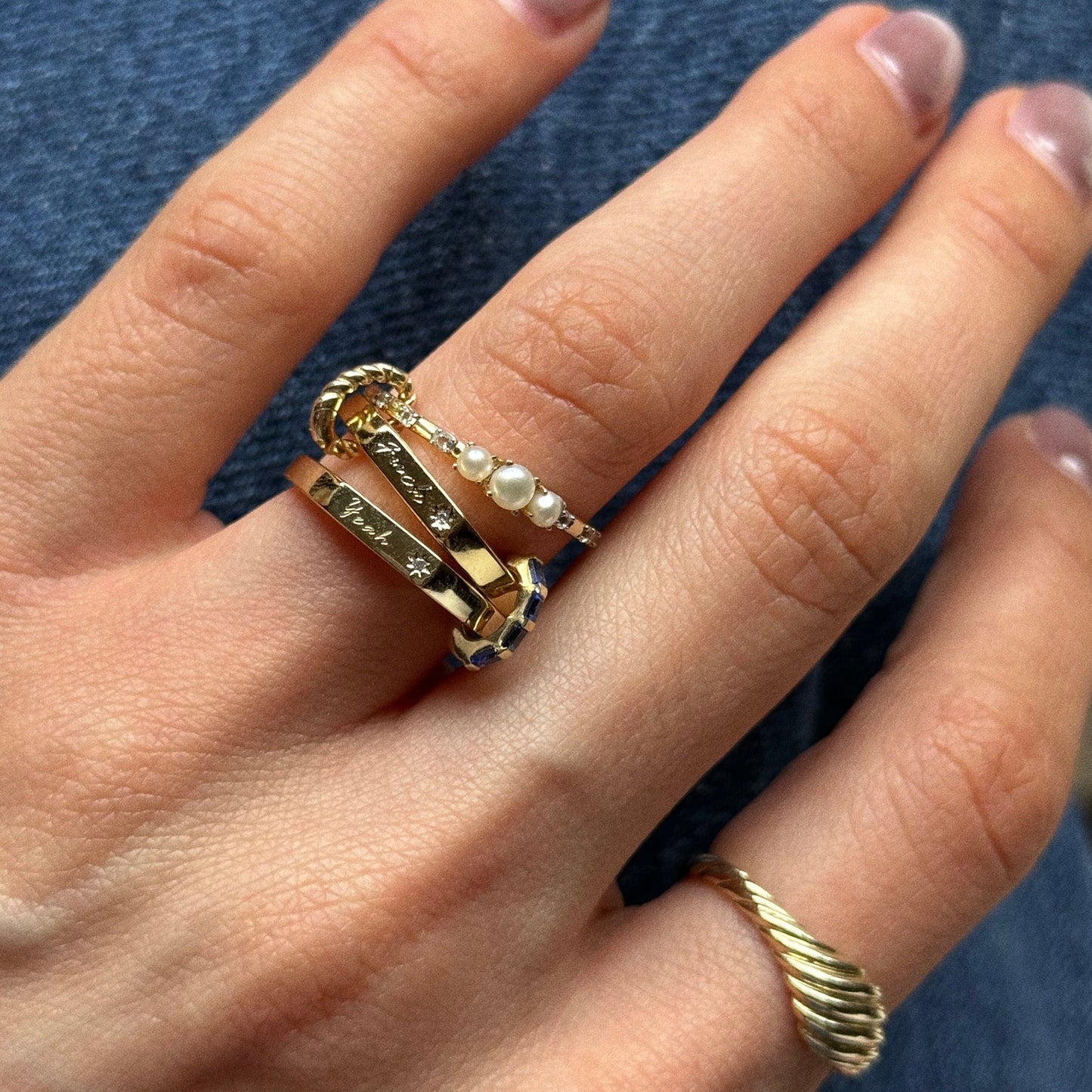 A hand with neatly manicured nails showcases the Ring Clip—stacked gold rings featuring engraved bands, a pearl-accented ring, and a twisted gold Ring Clip worn on the pinky finger.