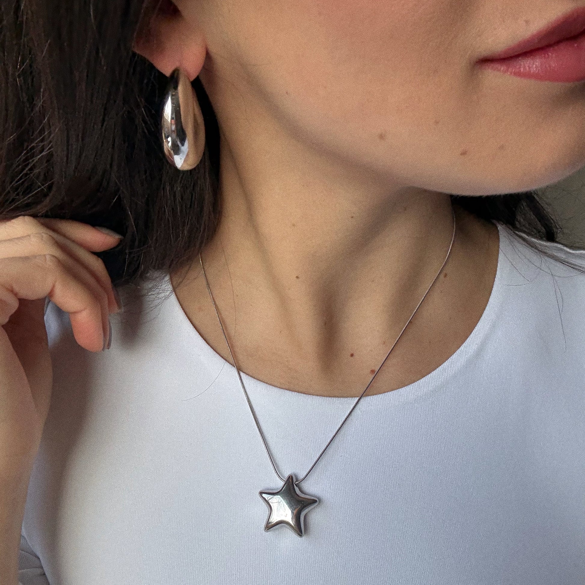A close-up of a woman with long dark hair wearing a white top, Casa Clara silver hoop earrings, and the Prism Necklace—a silver star-shaped pendant—while she touches her earring with one hand.