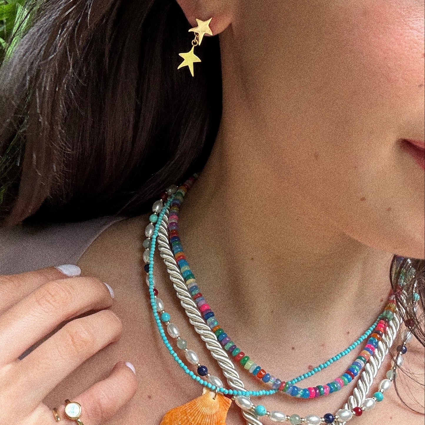 A woman wears colorful beaded necklaces, a shell pendant, and Star Drop Earrings. She also dons a gold ring with a white stone, her nails are painted white, and her dark hair frames her face.