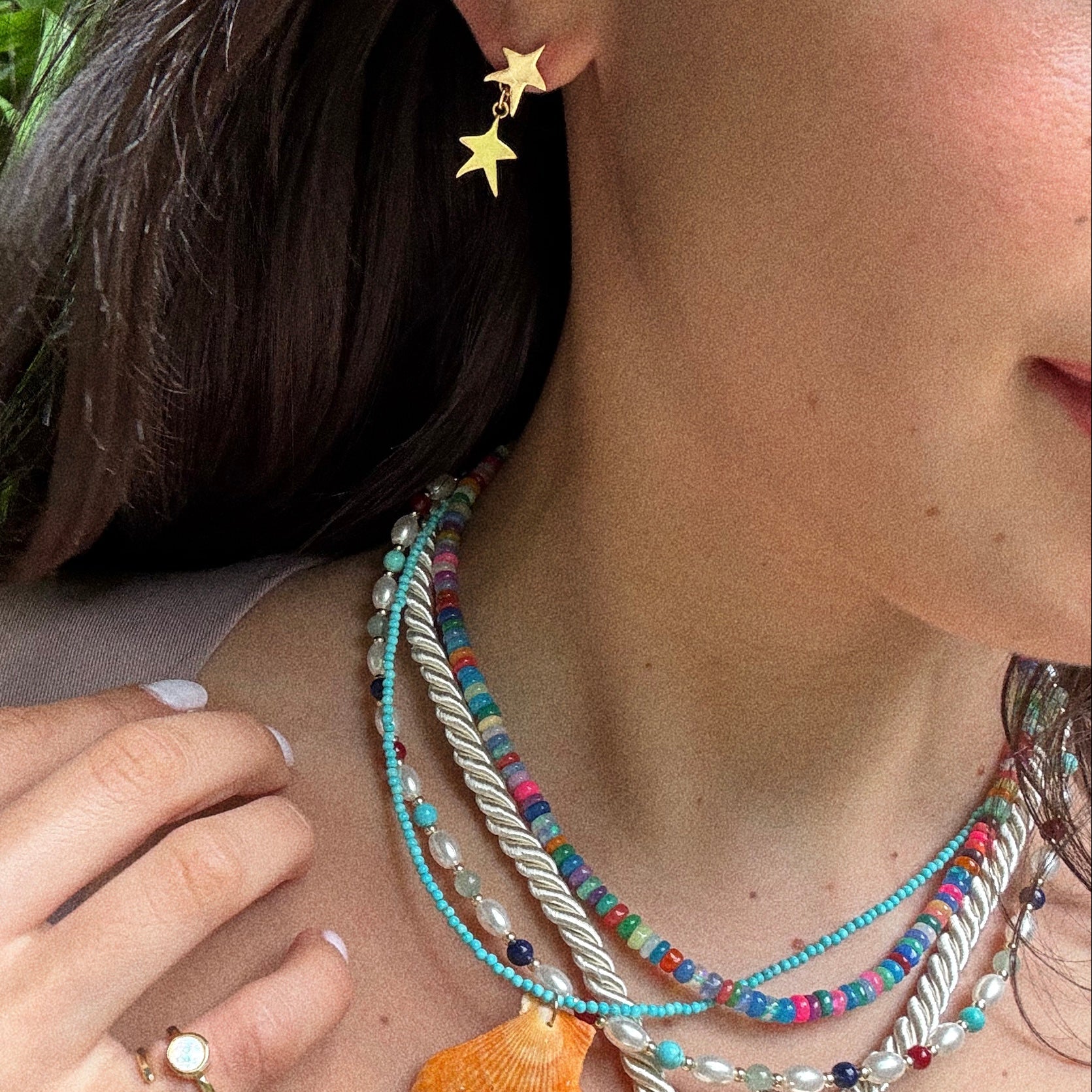 A woman wears colorful beaded necklaces, a shell pendant, and Star Drop Earrings. She also dons a gold ring with a white stone, her nails are painted white, and her dark hair frames her face.
