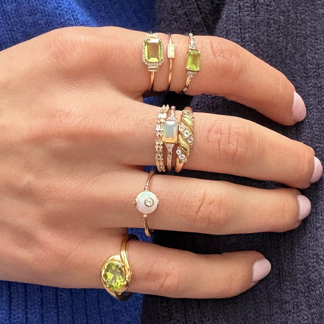 A hand with light pink nails wears eight gold rings, including the Solid Gold Glimmering Birthstone Baguette Ring featuring green, white topaz, and clear stones, all set against a blue textured fabric background.