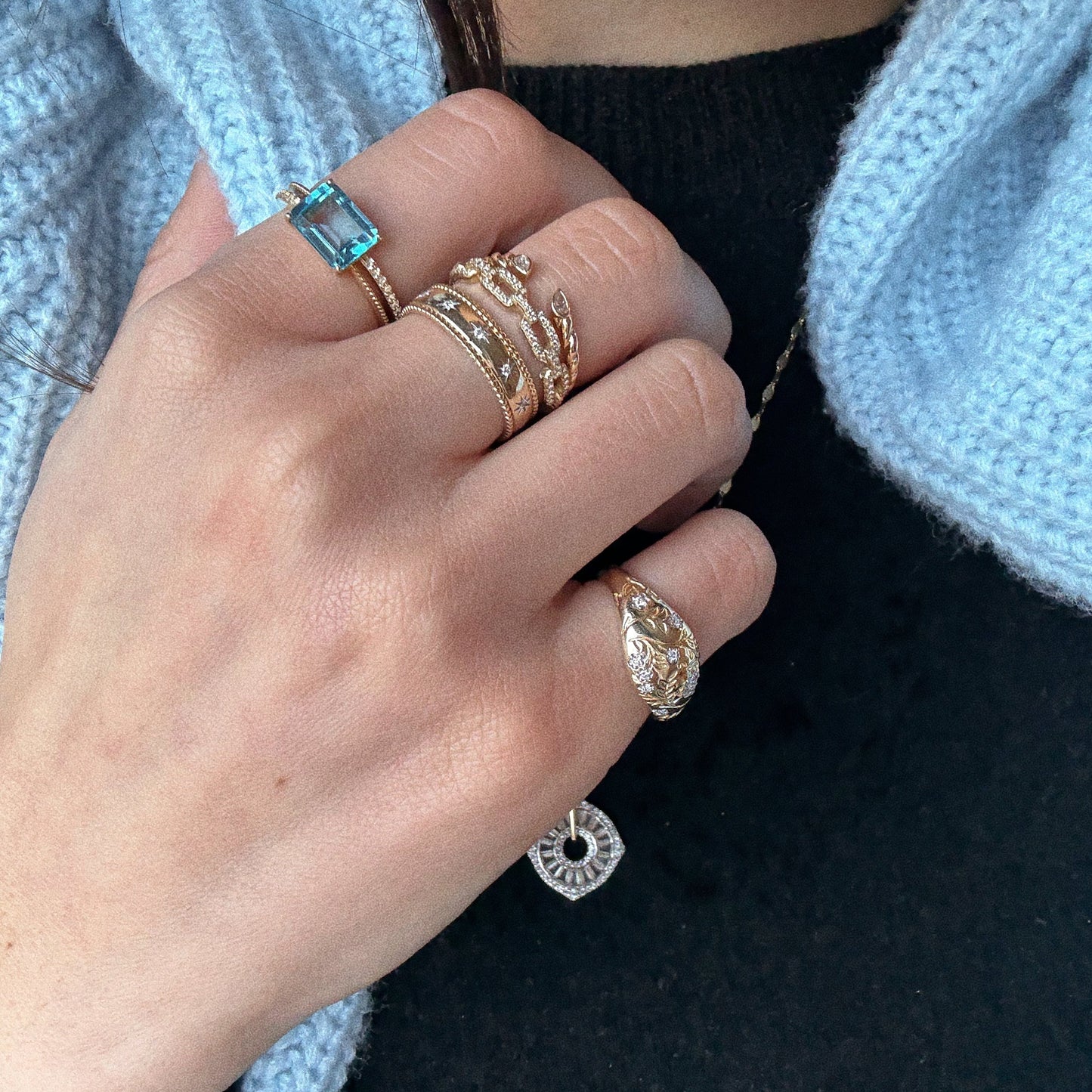 A hand wearing a 14K Blue Topaz Ring (Sample Size 7) shines with intricate gold designs, set against a black top and light blue knitted sweater.