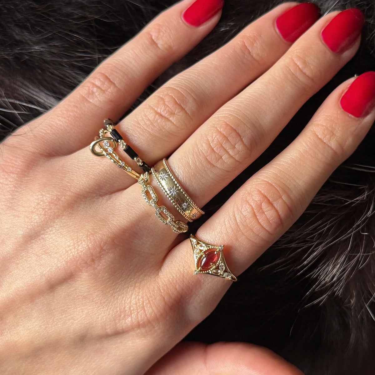 A hand with red nail polish wears three rings: a gold leaf band on the index finger, a silver band on the middle finger, and the 2026 January Capsule Art Deco Garnet Ring—ideal for January birthdays—on the ring finger, against dark fur.