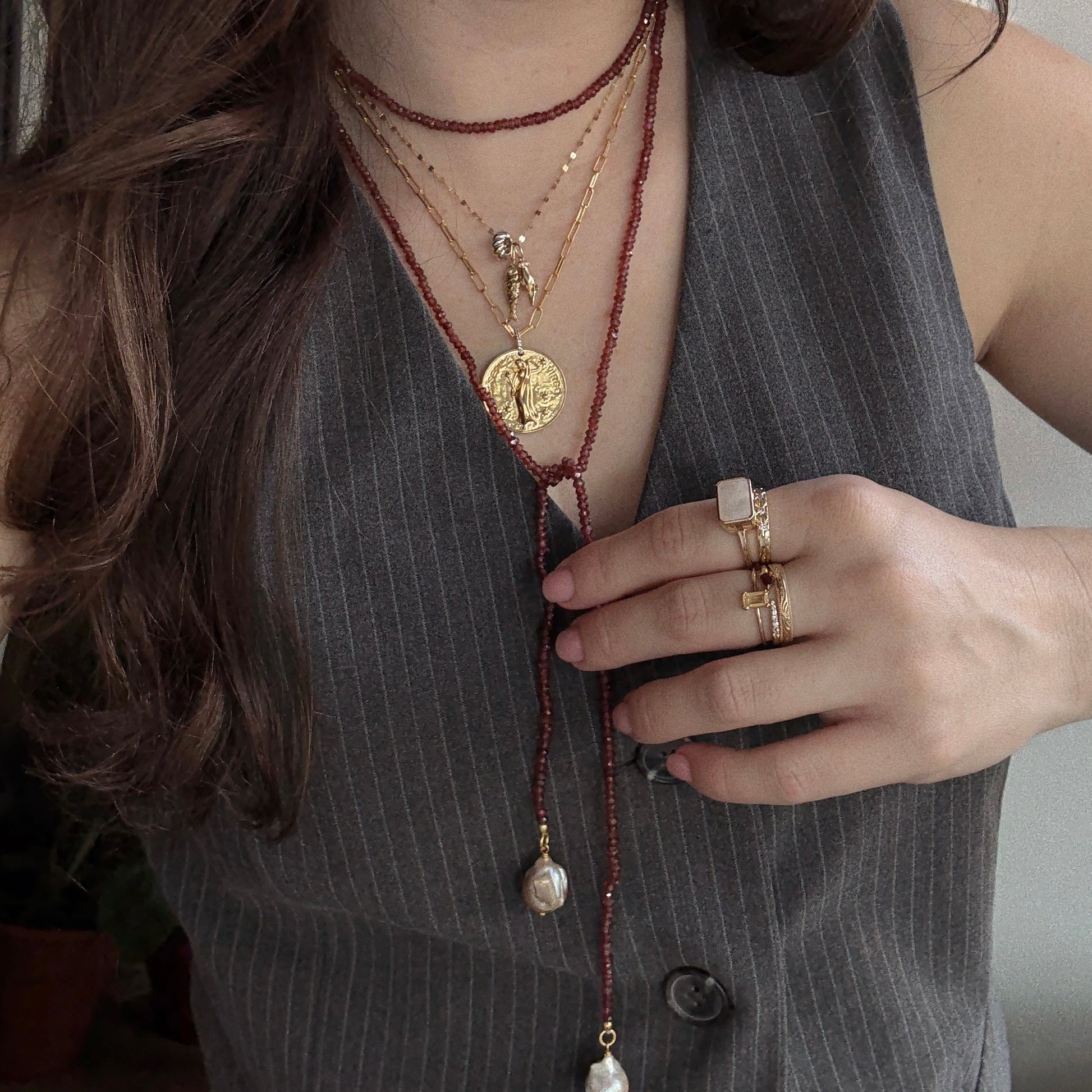 A person in a pinstripe vest layers gold necklaces, including the Gemstone Tie Necklace and a beaded pearl strand, with multiple gemstone gold rings and wavy hair draped over one shoulder.