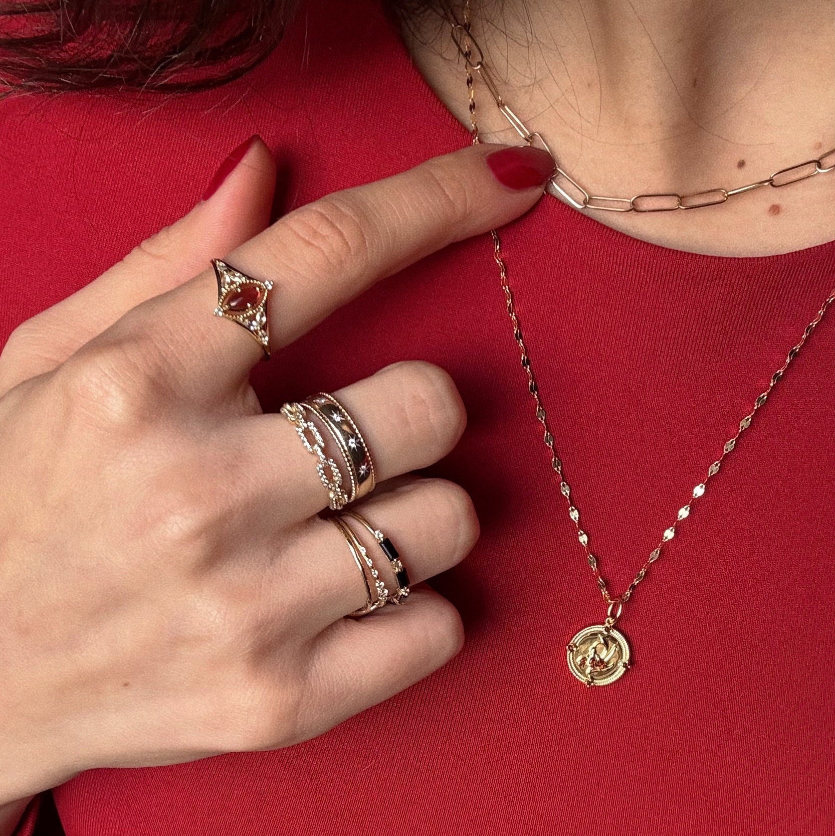 A hand with red nails, wearing the Solid Gold 2026 January Capsule Art Deco Garnet Ring and other gemstone rings, touches a gold chain necklace. Another gold necklace with a round pendant lies on a red top.