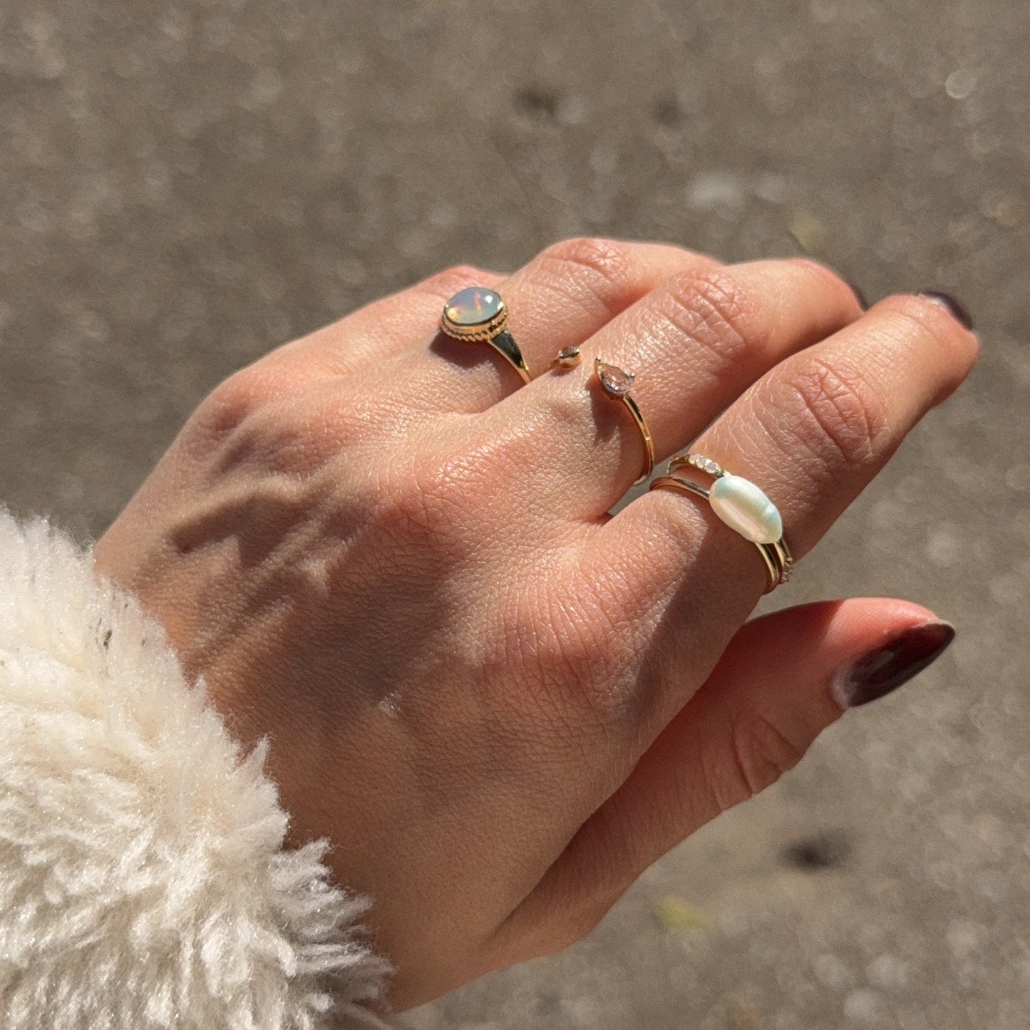 A hand with manicured nails wears the Solid Gold Textured Opal Signet Ring—featuring a delicate 1 mm gold band and an opal. The background is blurred, and the person wears a soft, cream-colored fuzzy sleeve.