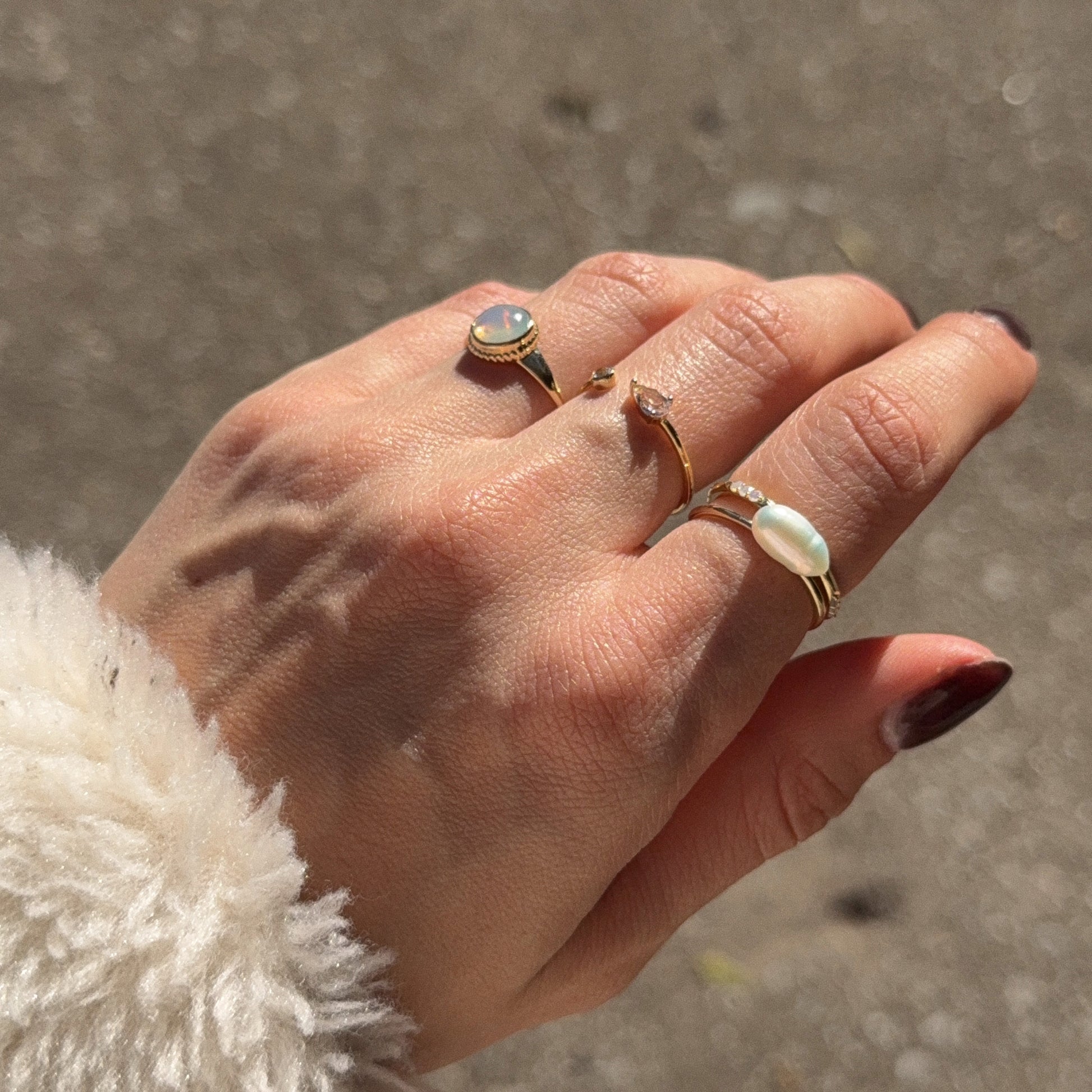A hand with manicured nails wears the Solid Gold Textured Opal Signet Ring—featuring a delicate 1 mm gold band and an opal. The background is blurred, and the person wears a soft, cream-colored fuzzy sleeve.