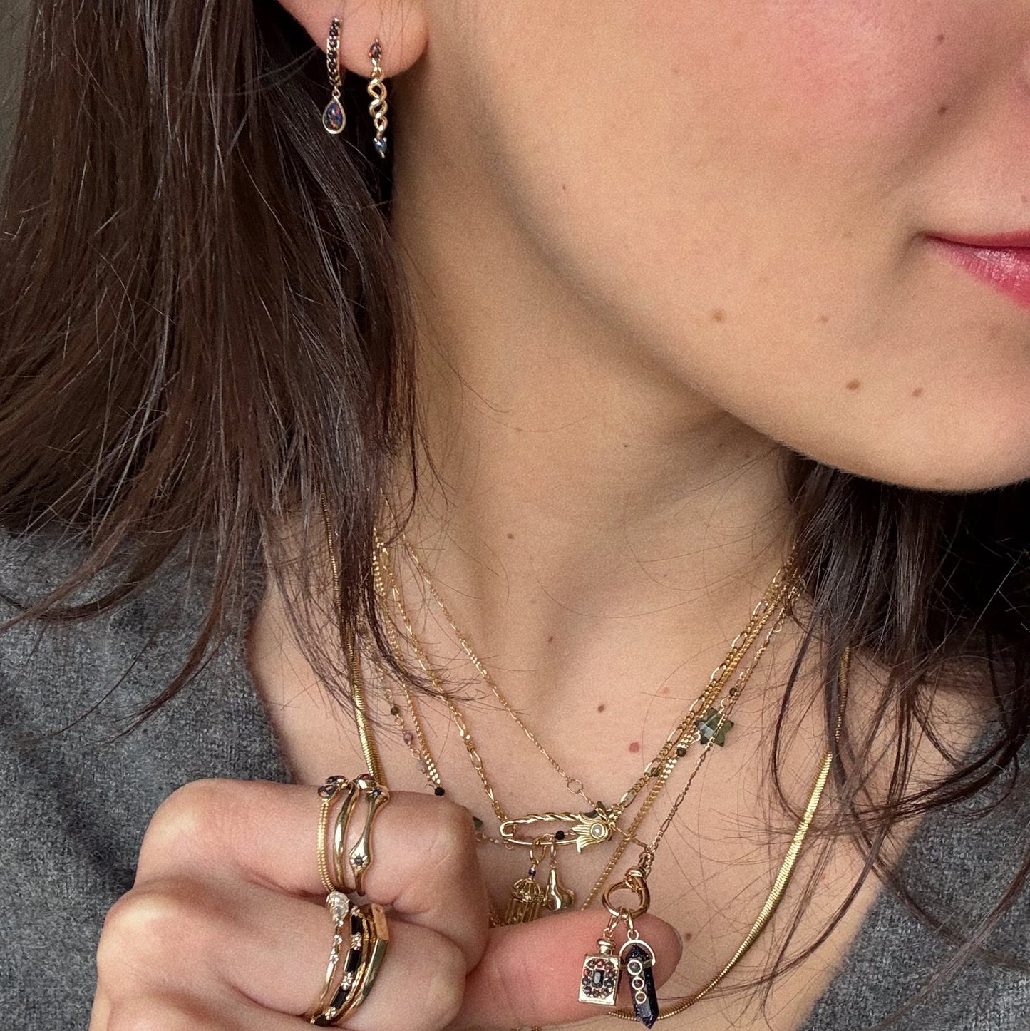 A close-up of a woman with dark brown hair wearing layered gold necklaces, gold rings, and Solid Gold Black Opal Galaxy Droplet Huggie Hoops. She holds her hand near her chin.