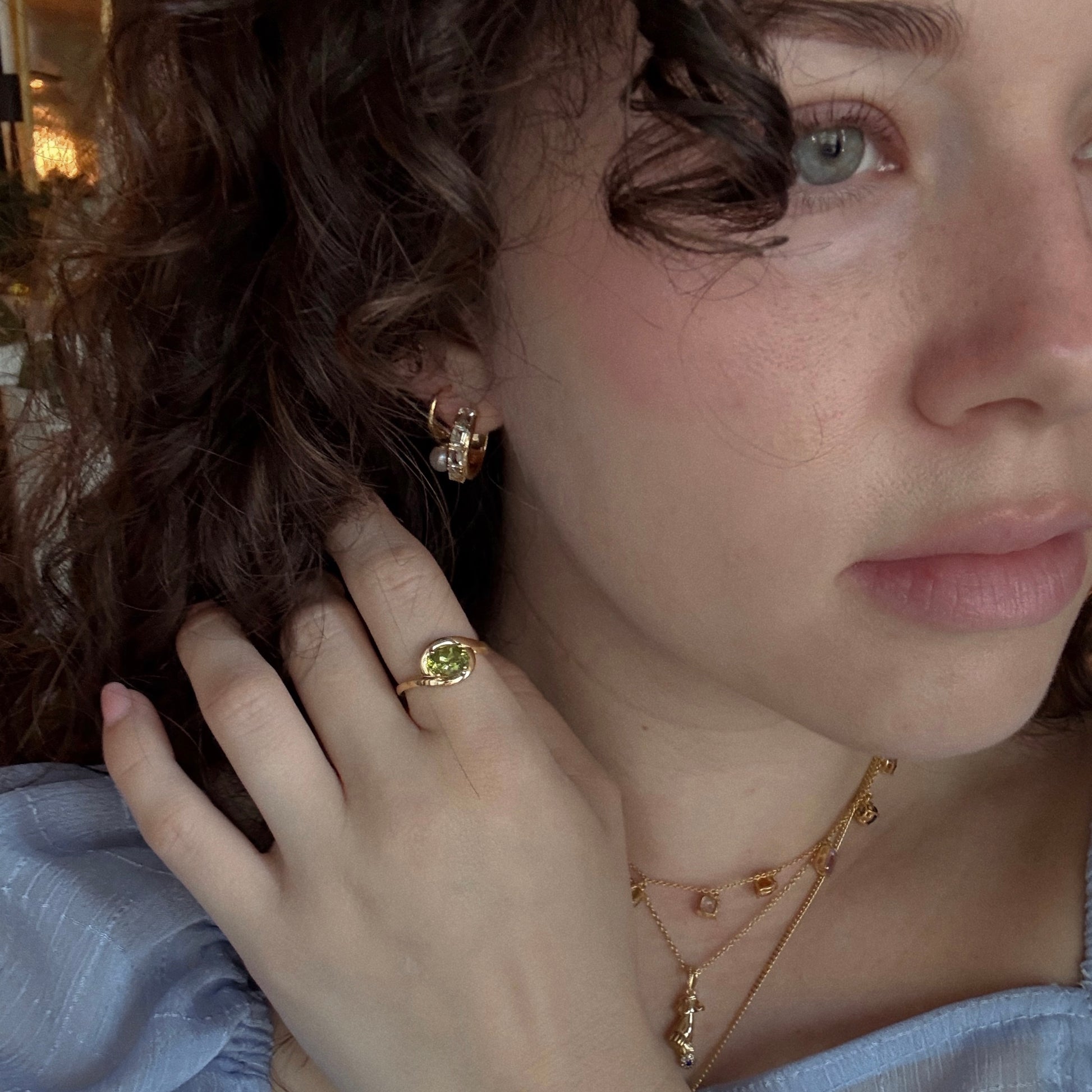 A woman with curly hair wearing a blue dress and gold jewelry, including Green Amethyst Era Huggie Hoops.