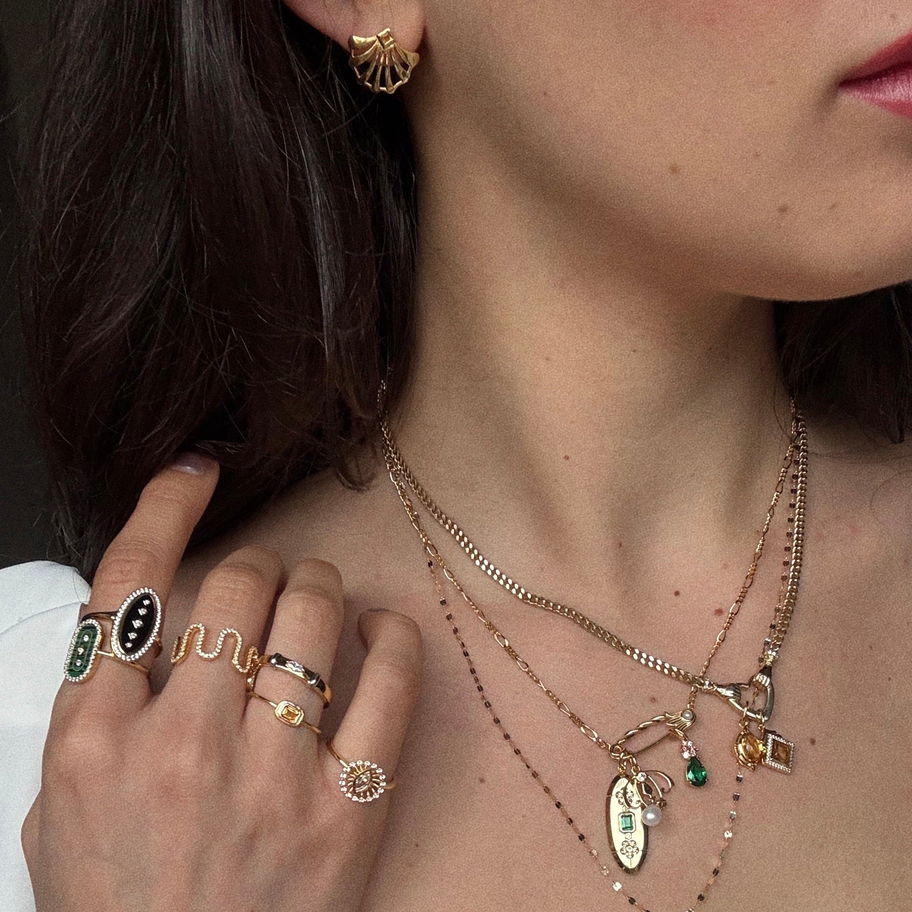 A woman’s hand touches her neck, displaying gemstone rings and 14k yellow gold vermeil necklaces. She wears the Showgirl Citrine Fan Earring, a gold seashell-inspired piece, alongside pendants in green, black, citrine, and gold tones.