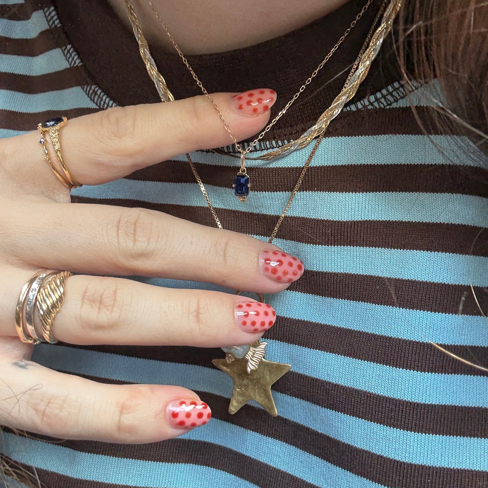 A hand with red and pink polka dot nails holds a solid gold necklace. Multiple gold rings and layered necklaces, including the Solid Gold 2025 September Capsule Classic Blue Sapphire Pendant Necklace, are worn over a brown and blue striped shirt.