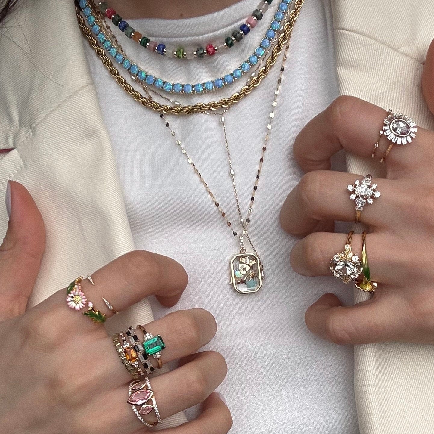 A person wears a cream blazer and white top, layered with vibrant beaded necklaces and the Solid Gold Tiny Lucky Charm for Glass Charm Locket, plus multiple gemstone and floral rings, displaying detailed, colorful jewelry.