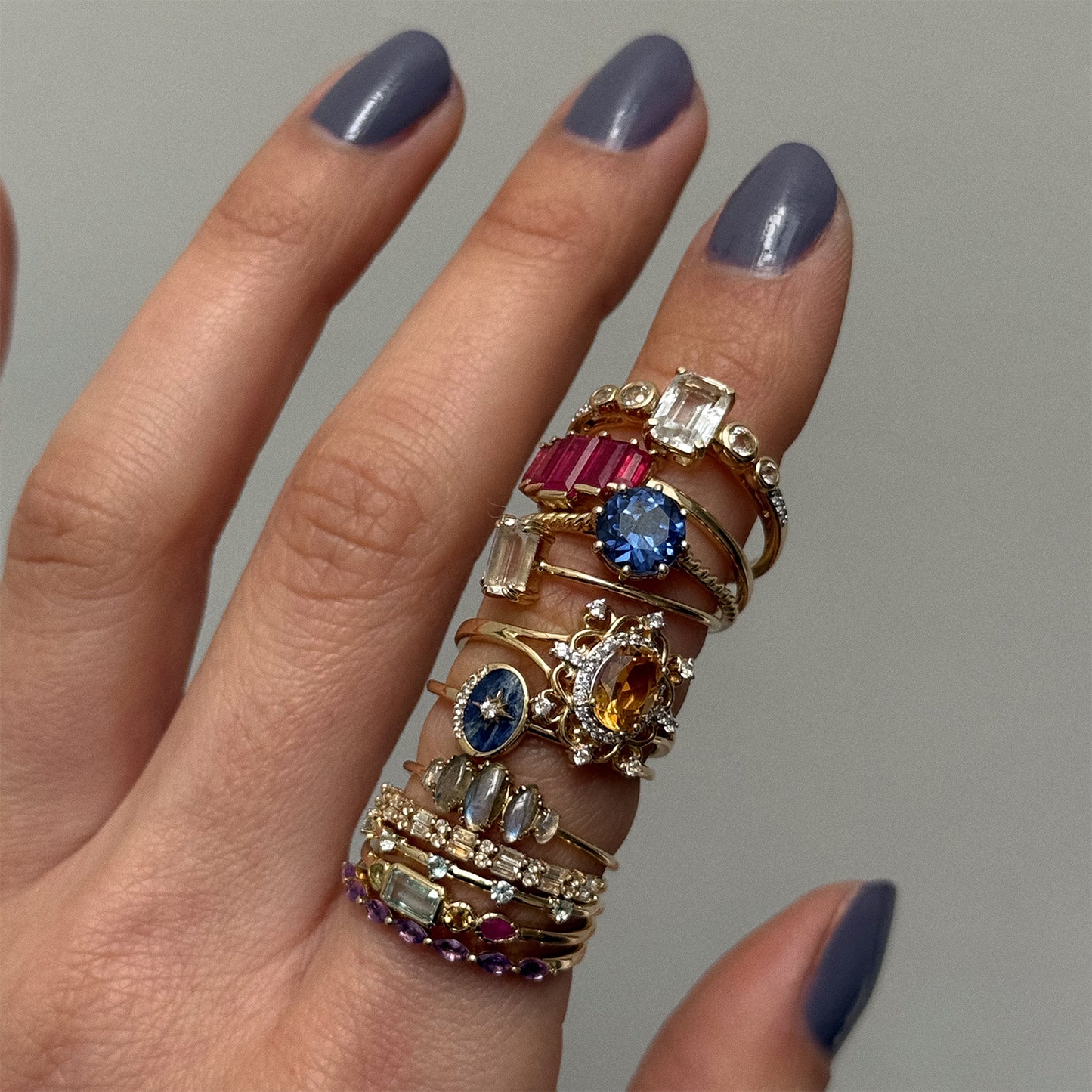 A hand with purple nail polish displays the Solid Gold Surprise Ring plus vibrant gemstone rings in blue, red, clear, and yellow, all stacked stylishly on each finger in unique shapes and sizes.