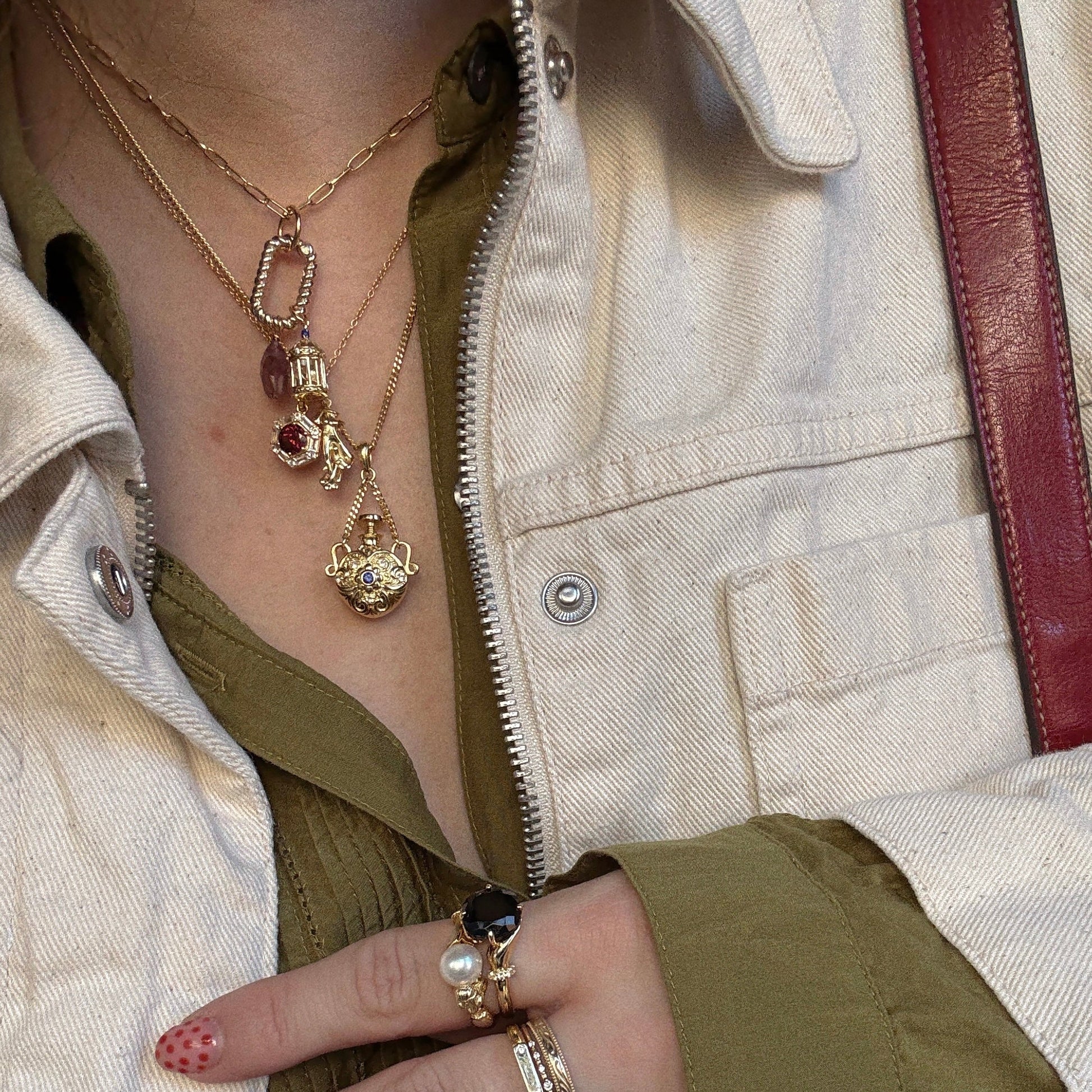 A person wears layered, vintage-inspired gold necklaces—including the Juliet Vial Pendant Necklace and a blue sapphire piece—paired with a cream jacket, olive shirt, rings, and nails painted with delicate red polka dots.