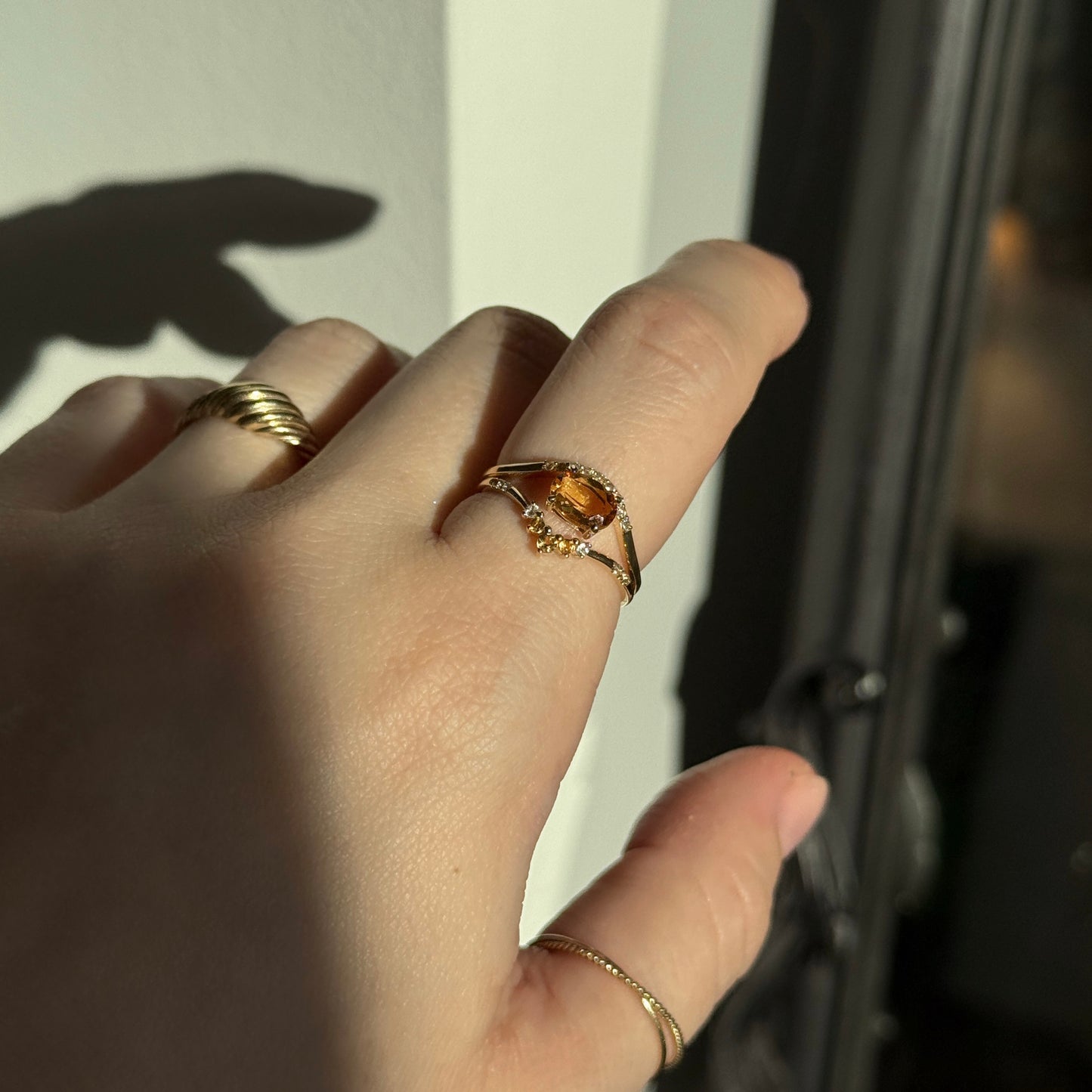 A hand adorned with the Solid Gold 2025 November Capsule Citrine Ring Set with Free Gift—November’s birthstone jewelry—gleams in sunlight by a dark window, casting an elegant shadow on the wall.
