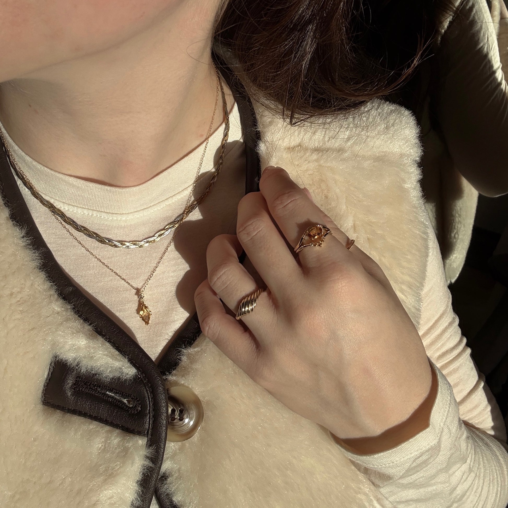 A person wears the Solid Gold 2025 November Capsule Citrine Statement Set with two necklaces and two rings, paired with a cream top and beige vest, as sunlight casts shadows across their hand and chest. Free gift included.