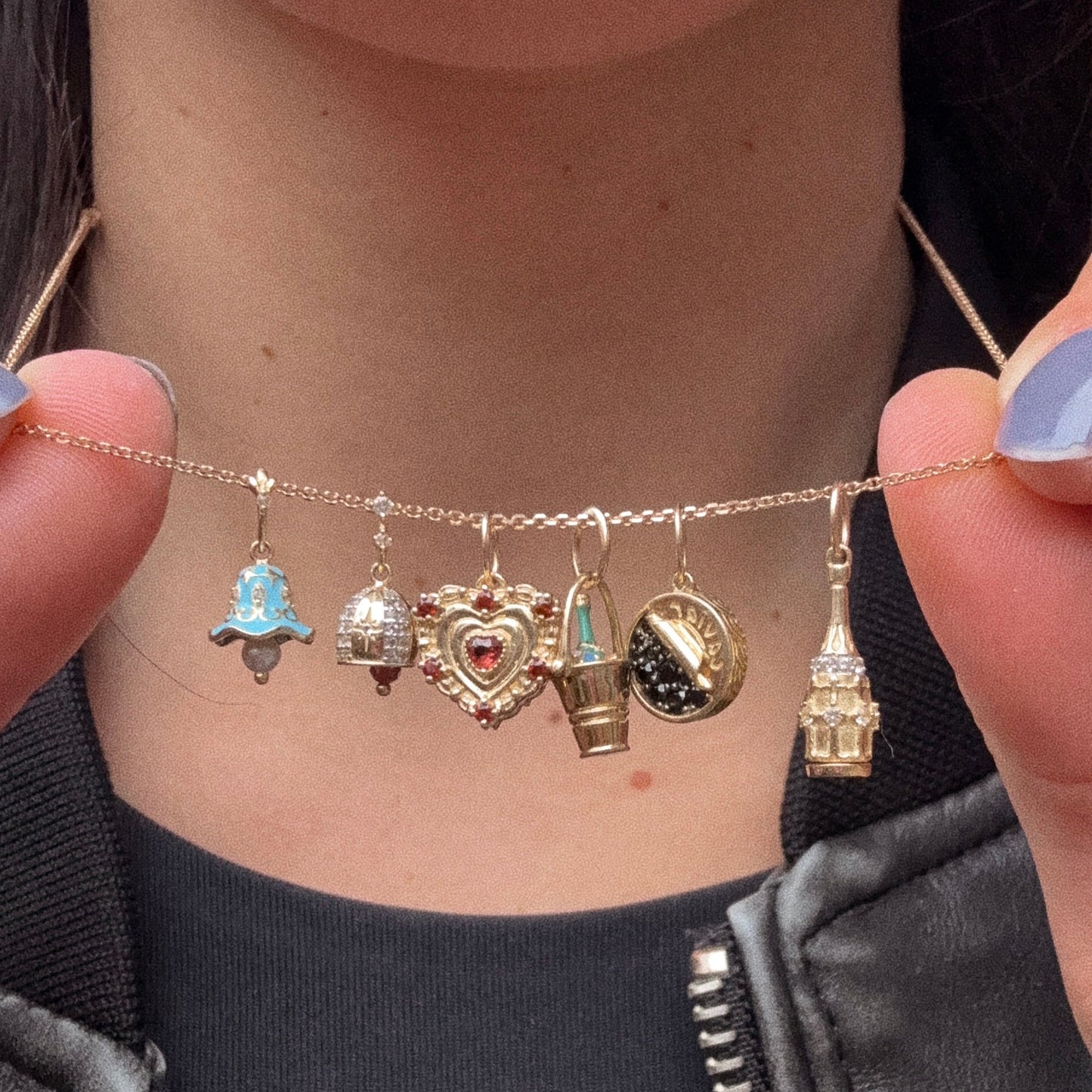 A person holds the Solid Gold Celebration Charm necklace, crafted in 9k solid yellow gold, featuring six unique charms—including an engraved charm, blue bell, silver bell, heart, turquoise cone, and gold building—against a black shirt and jacket.